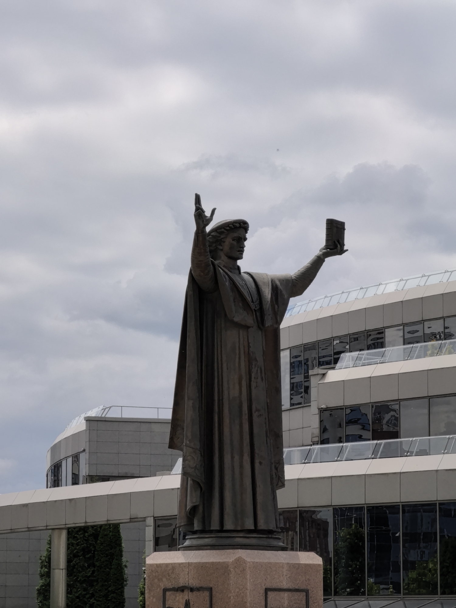 A bronze statue of a person stands prominently in the foreground against a cloudy sky, with modern buildings in the background. The statue depicts a figure wearing a robe and a laurel wreath, holding a book in their outstretched right hand and with their left hand raised with two fingers extended. The statue is on a stone pedestal. Behind the statue, a multi-story building with large glass windows and balconies is visible. The windows reflect the surrounding environment and the sky. Green trees are also visible behind the pedestal. The overall scene suggests an outdoor public space, likely in a city.
