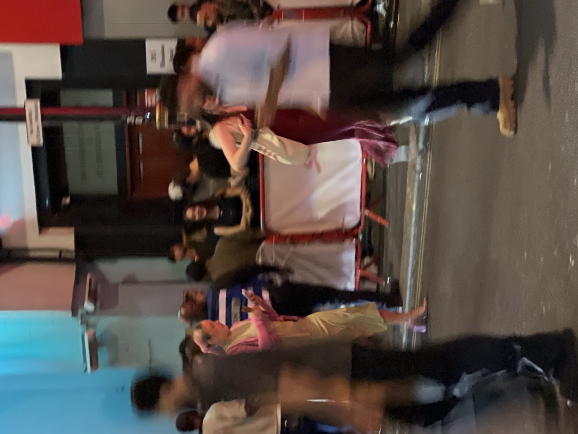 The image depicts a bustling street scene, likely in London, United Kingdom, as suggested by the architectural style and general atmosphere. The photo is somewhat blurry, indicating motion or a low shutter speed.
Numerous people are visible, some standing, others walking. Clothing styles vary, suggesting a diverse crowd. People appear to be socializing and moving along the pavement.
A temporary barrier, perhaps for crowd control, is visible near the center of the frame. The background includes building facades with signage and architectural details typical of urban areas in London. The pavement is dark, suggesting either the time of day or wet conditions.