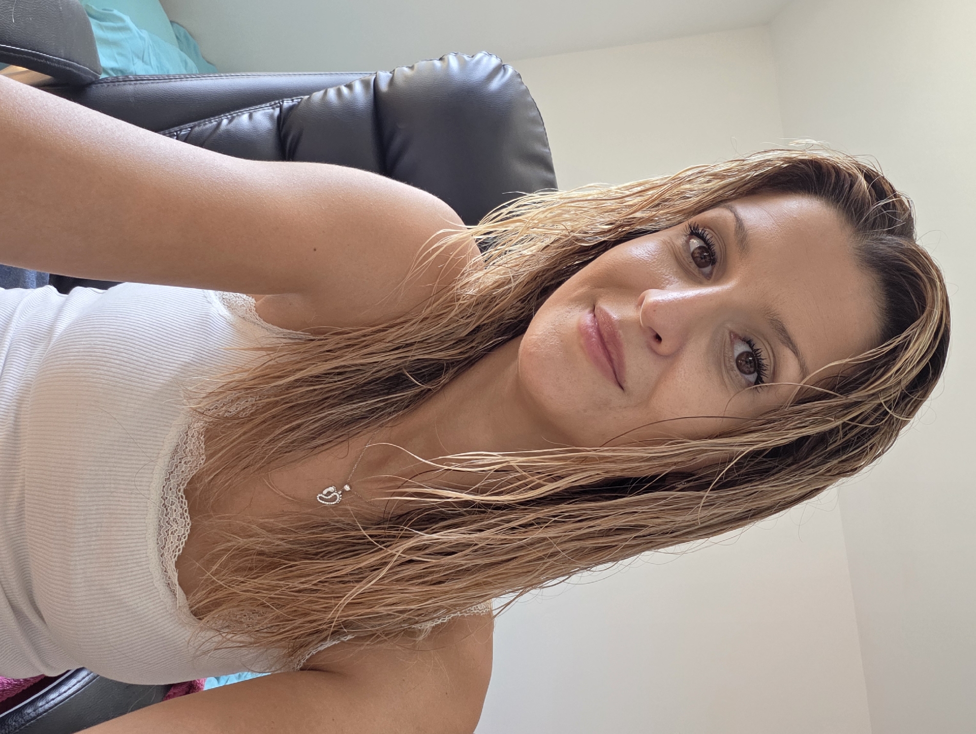 A medium shot selfie shows a woman with wet, blonde hair looking directly at the camera. She is wearing a white tank top with lace trim and a necklace with a footprint pendant. She is seated in a black office chair in front of a white wall. The lighting suggests an indoor setting, possibly a home office. The background is minimally detailed, with a portion of a blue and white striped bedding visible to the left. No specific location in Munich, Germany is identifiable from the visual cues in the image.