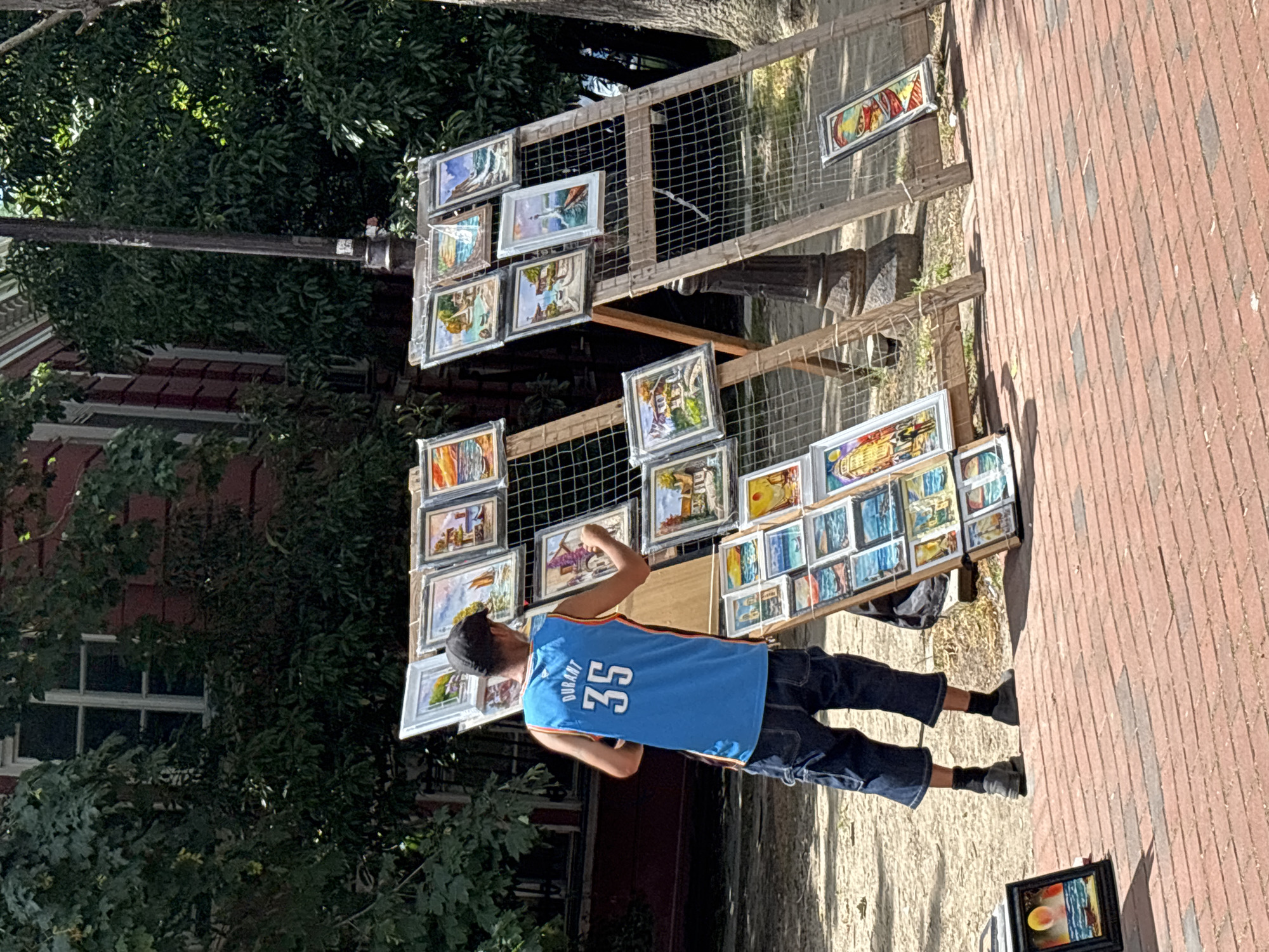 The image shows a person displaying paintings for sale on a wooden stand outdoors. The person, wearing a blue basketball jersey with "DURANT 35" visible on the back and dark pants, appears to be arranging or presenting the artwork. The paintings are displayed on a multi-tiered wooden rack, with many of them showing landscapes and seascapes. The setting is a brick-paved walkway, suggesting a public area or street market. Trees and a building with a window are visible in the background, partially obscuring the view. The lighting suggests it is daytime, likely sunny, given the shadows cast on the ground. The overall scene depicts a street vendor selling art. No specific emotions are discernible. The location is identified as Varna, Bulgaria.