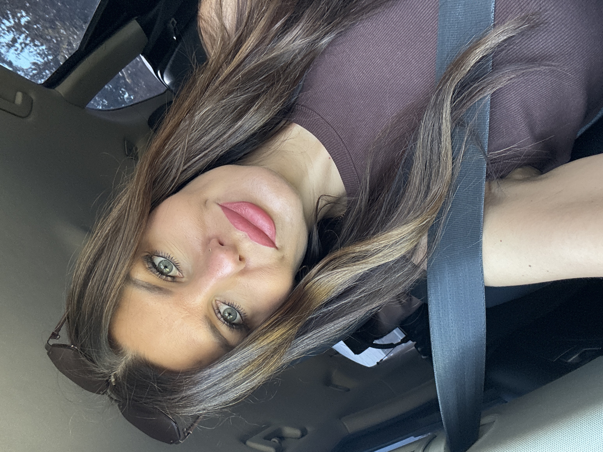 A woman is taking a selfie in a car. She has long, brown hair with blonde highlights and green eyes. She is wearing a brown top and sunglasses on her head. A seatbelt is visible across her chest. The background shows the interior of a car and a view of trees through the window. The image is taken from a slightly high angle, looking down at the woman. The location is not specified in the image, and therefore cannot be identified as Noviy, Russia.