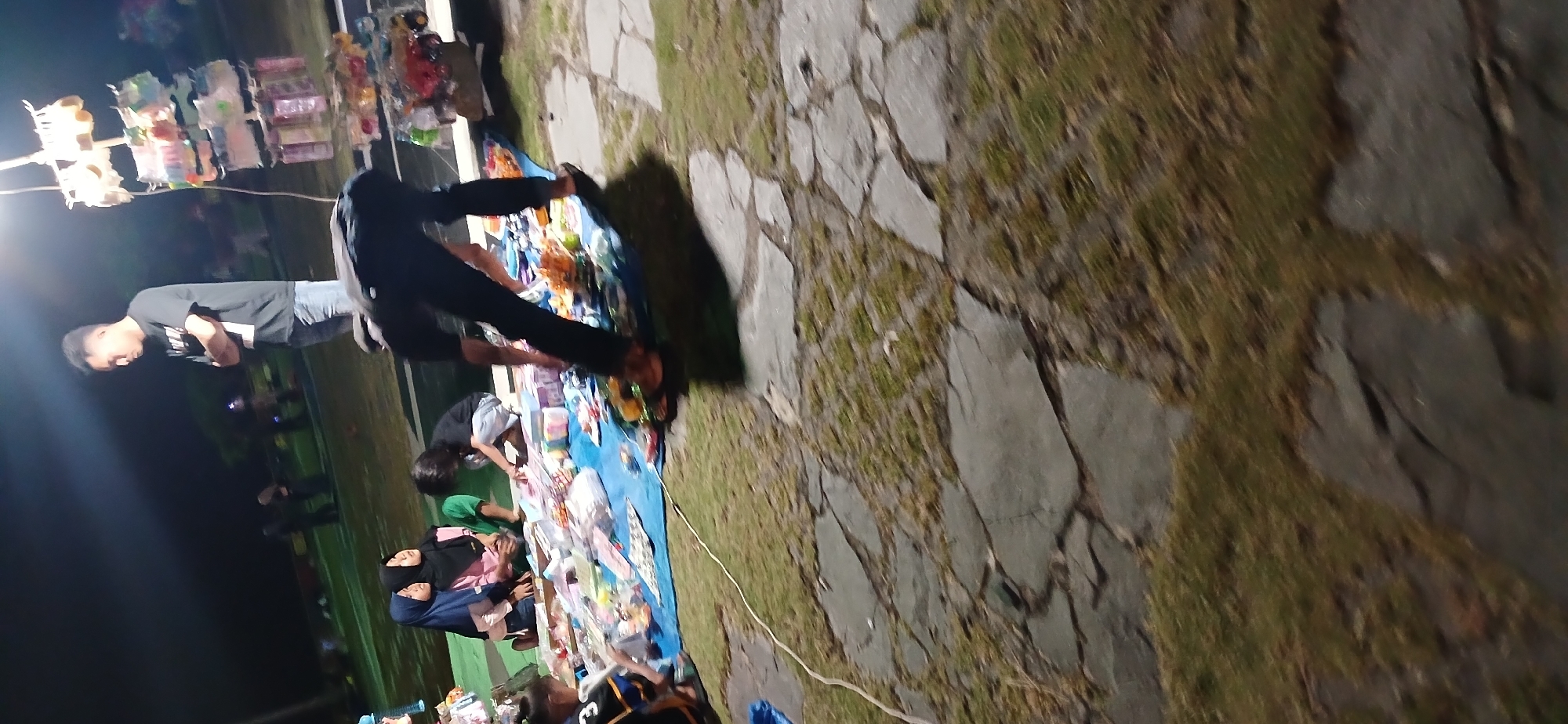 In Tanjung Balai Karimun, Indonesia, at night, a group of people are gathered outdoors, possibly at a market or event. A man in a black t-shirt and jeans stands with his arms crossed, looking down. Another person, whose face is obscured, is bending over a blue tarp laid out on the ground. This tarp is covered with various items, possibly merchandise for sale, including colorful plastic containers and small toys. Several individuals, including two women wearing hijabs and a child, are seated or kneeling around the tarp, interacting with the items. In the background, there are other people and illuminated stalls, suggesting a lively atmosphere. The ground is a mix of grass and large, flat stone slabs.