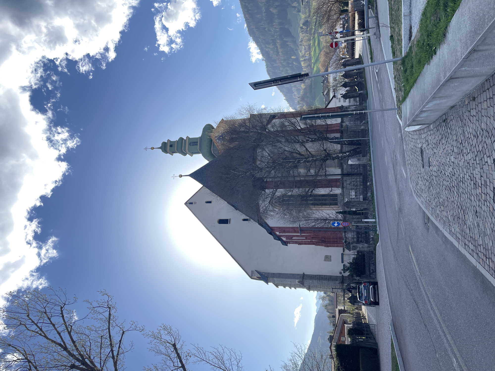 The image shows the church of St. Margaret in Sterzing-Vipiteno, Italy, from a low-angle perspective looking up. The church features a tall, green-roofed steeple and a white facade with red architectural details. The sun is positioned behind the building, causing a glare. A road runs alongside the church, with a few cars parked on it. A sidewalk lined with stone pavers is visible on the right. Bare tree branches are in the lower left corner and partially obscuring the church. The sky is blue with puffy white clouds. In the background, a hillside and buildings are visible. A traffic sign is on the side of the road.