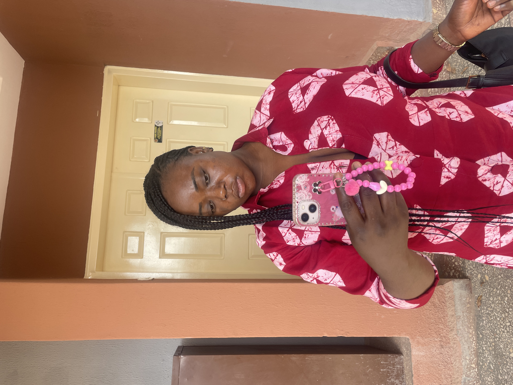A woman with braided hair and a nose piercing is standing in front of a light yellow door. She is holding a pink smartphone with a beaded charm. The woman is wearing a red patterned dress and a gold watch. The background consists of a peach-colored wall on the left, a gray concrete-like surface on the right, and a glimpse of a black bag. The location is Jos, Nigeria.