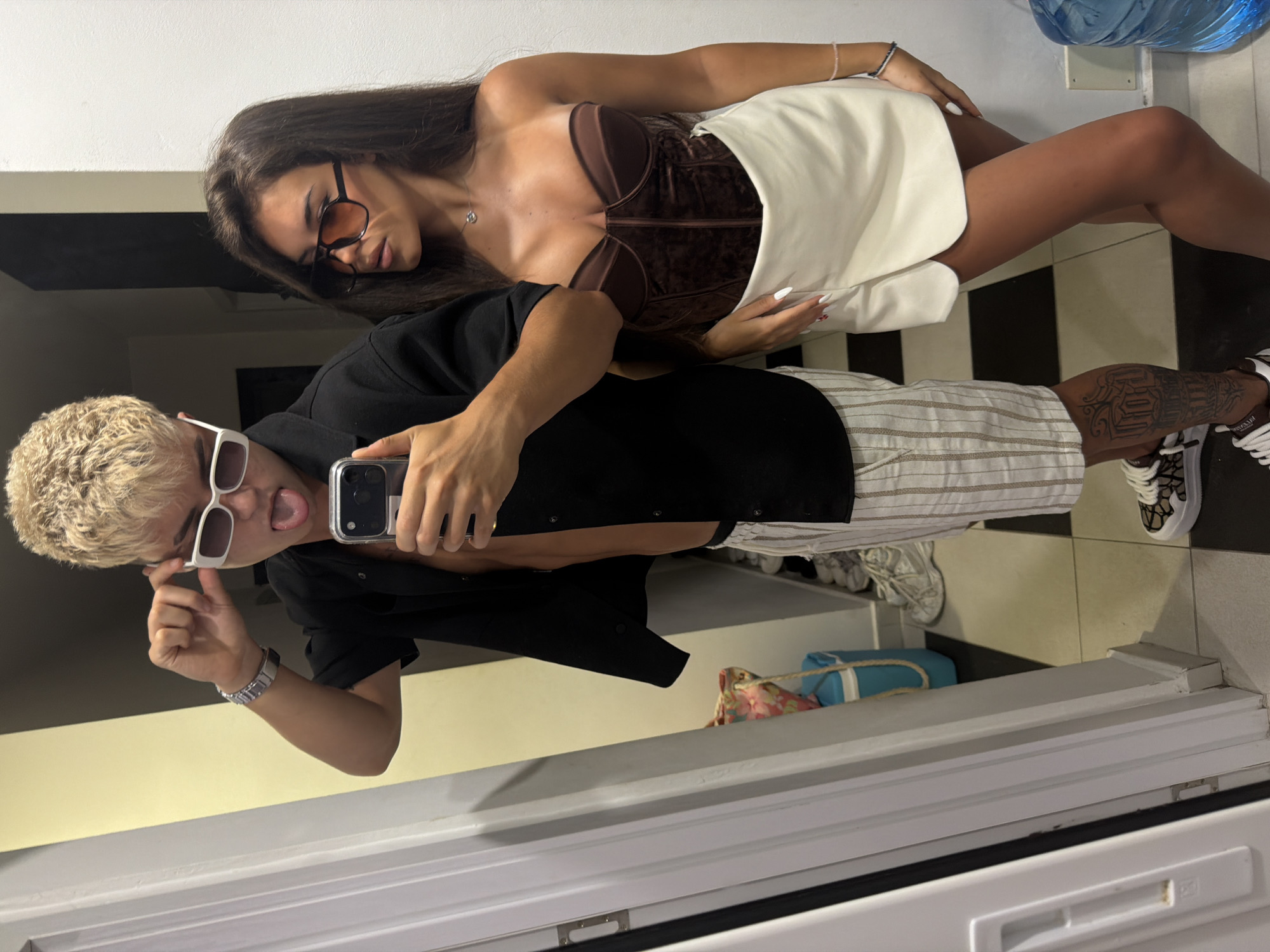 In a full-length mirror selfie, a young person with blonde, spiky hair and white-rimmed sunglasses playfully sticks their tongue out. They are wearing a black open-collared shirt, revealing a portion of their torso, and vertically striped shorts. To their right stands a young woman with long, dark brown hair, wearing large, orange-tinted sunglasses. She is dressed in a brown velvet corset top and a white, skort-like skirt. They are posing together in what appears to be an indoor setting with a tiled floor and a white wall. A blue cooler bag and other items are visible in the background, reflected in the mirror. The overall scene suggests a casual, possibly pre-outing moment captured for social media.