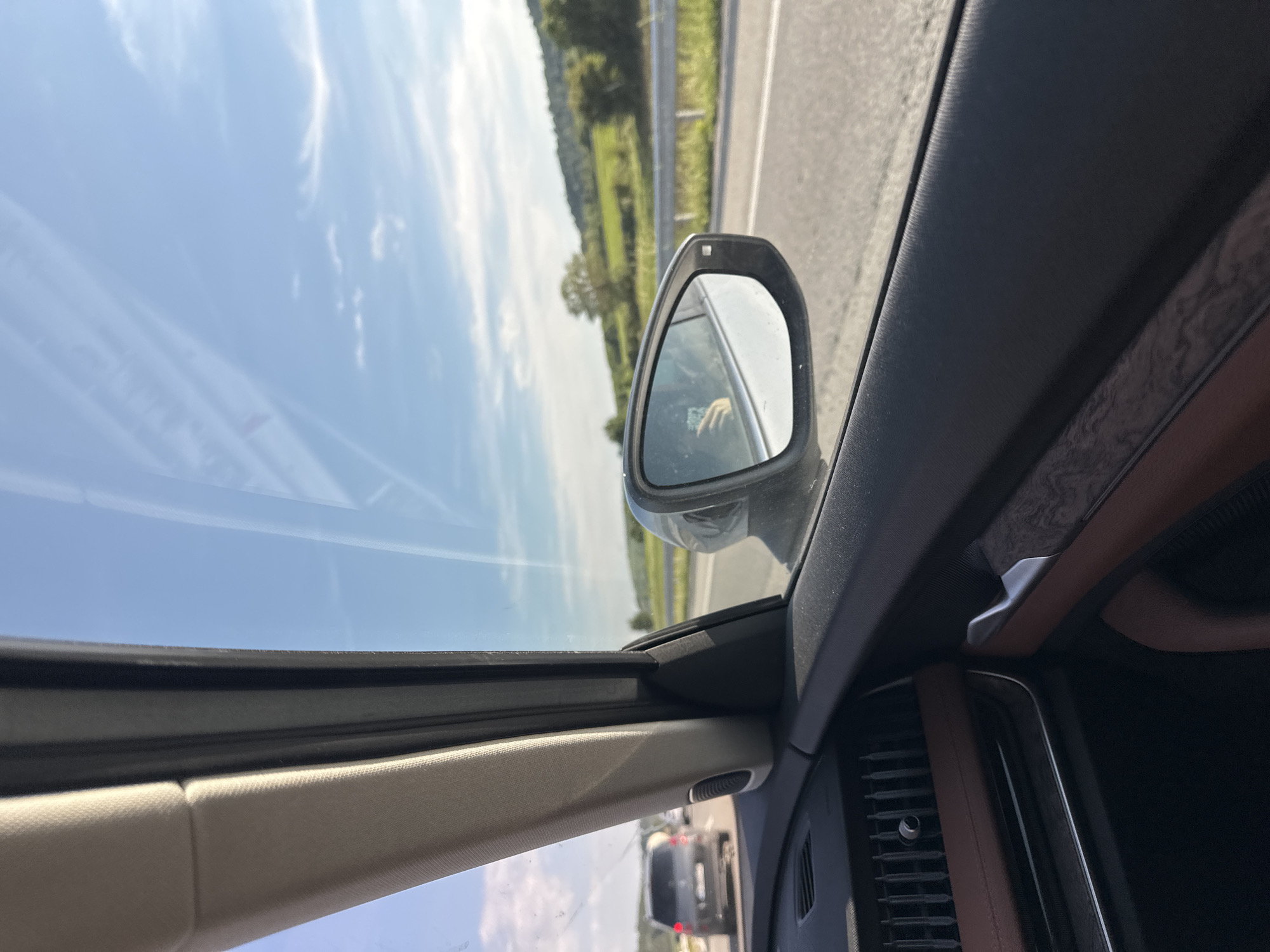 The image depicts a daytime scene from the interior of a moving vehicle, looking outward through the passenger side window onto a highway. The location is Dren, Bulgaria.
In the immediate foreground, the interior of the vehicle is visible, featuring a beige fabric-covered A-pillar extending vertically from the lower left to the upper center. Below the A-pillar, a dark dashboard with a horizontal air vent and a brown trim element is present. The dark door panel, also with a brown trim accent and a metallic door handle, is visible in the lower right quadrant.
The passenger-side rearview mirror is centrally positioned on the right edge of the frame. Within the mirror's reflection, a person's left hand is visible holding a dark-colored smartphone, indicating the photographer or a passenger is present and likely using the device.
Beyond the vehicle, a paved, multi-lane highway stretches into the distance. A metallic guardrail runs parallel to the highway on the right. In the far distance, a dark gray SUV with illuminated taillights is visible, suggesting traffic ahead. Other vehicles are partially visible further down the road.
The background consists of a natural landscape featuring rolling green hills and fields dotted with scattered trees under a clear, light blue sky. Sparse, wispy white clouds are present across the sky. The overall scene suggests a sunny day in a rural or semi-rural setting.