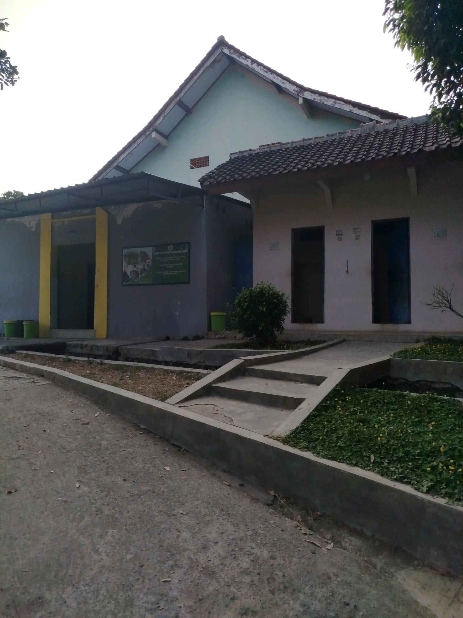 The image shows the exterior of a building that appears to be a public restroom facility, likely associated with a school or community center, given the sign that reads "SMP N 1 KARANGSAMBUNG". The building has a light green pitched roof with dark gray tiles, and a section with a flat dark gray roof extending over a portion of the front. The walls are painted in muted colors, with one section being a rough gray concrete and another section painted a light pinkish hue. There are two visible doorways on the pinkish section of the building, each with a dark blue door. In front of the building, a concrete walkway leads to the entrances, with steps and planters filled with green foliage and small yellow flowers. Green cylindrical trash bins are visible on the left side of the building. The ground in front is paved with asphalt, and a concrete curb runs along the walkway. Trees and foliage are present in the background and to the right, suggesting an outdoor setting. The sky is overcast and bright, indicating it is daytime. The overall scene is one of a functional, perhaps slightly aged, public building in an outdoor environment.