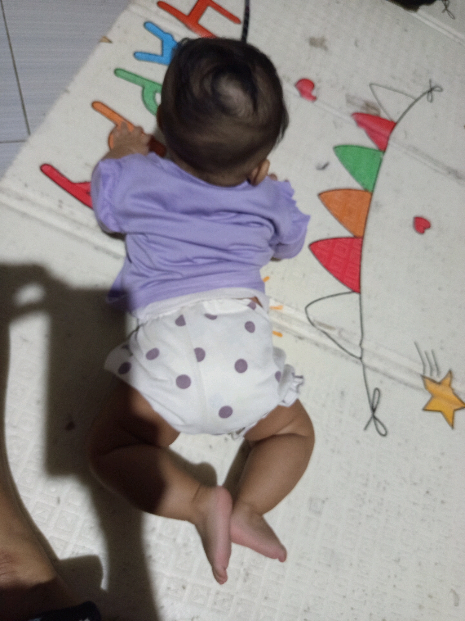 In Salebu, Indonesia, a baby is lying on their stomach on a playmat. The baby is wearing a purple shirt and a diaper with purple polka dots. The playmat has colorful letters and a drawing of a dragon. A person's leg is visible on the left side of the image, casting a shadow on the playmat.