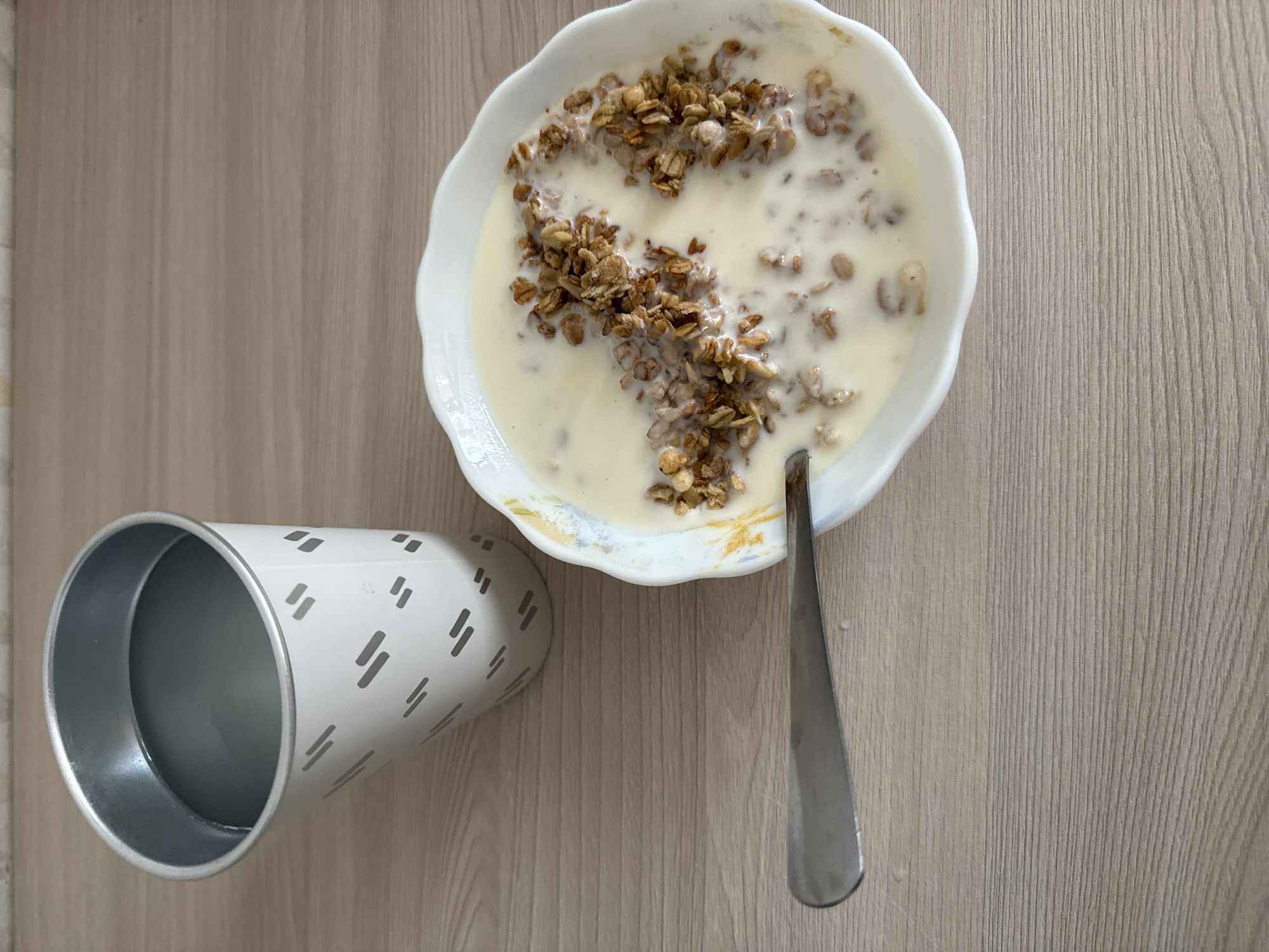 A bowl of granola with milk and a spoon are on a wooden table next to a cup of liquid. The bowl is white with a scalloped edge and is filled with a creamy liquid and granola. A silver spoon rests in the bowl, with its handle extending to the left. To the left of the bowl is a white cup with a pattern of gray dashes. The cup is partially filled with a clear liquid. The table is light brown with a wood grain texture.