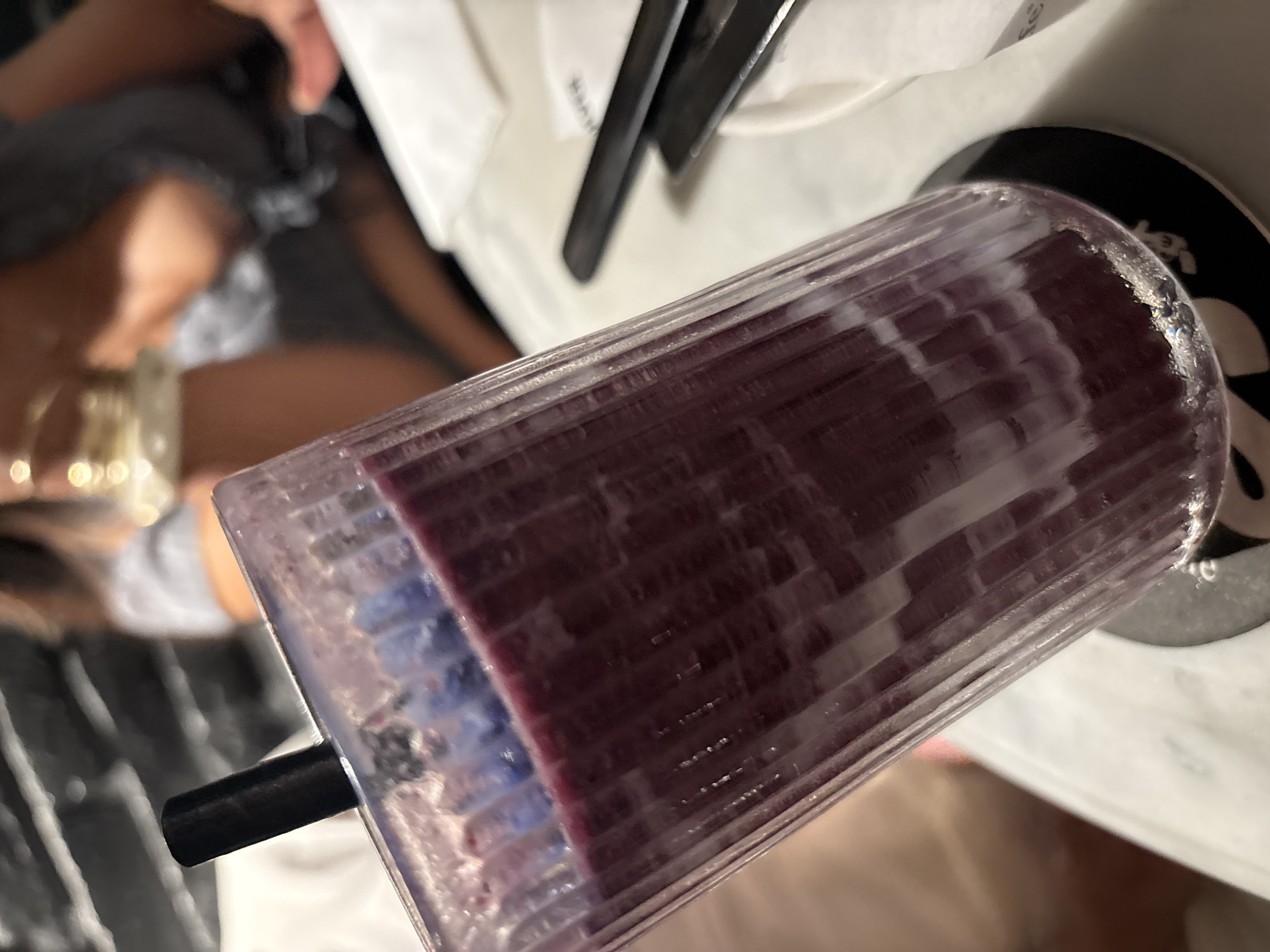 A close-up shot of a tall, fluted glass filled with a dark purple smoothie sits on a black and white coaster. A black straw is inserted into the drink. The glass is positioned on a white marble table. In the blurred background, a person is visible wearing a dark grey t-shirt, and another person is partially visible in a white shirt. The setting appears to be a restaurant or cafe in North Kuta, Indonesia. There are also chopsticks on a white napkin to the right of the glass.