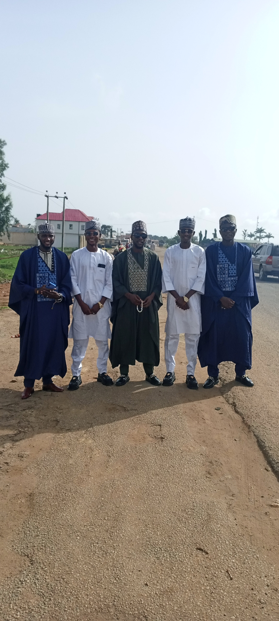 Five men are standing in a line on a dirt road in Naraguta Hausawa, Nigeria. They are all wearing traditional Hausa clothing and hats. From left to right, the first and last men are wearing dark blue robes over their outfits; the middle man is wearing a dark green robe over his outfit; and the two men standing next to him are wearing white outfits. The man in the middle is holding a string of beads. A building and electrical poles are visible in the background. A silver car is visible to the right.