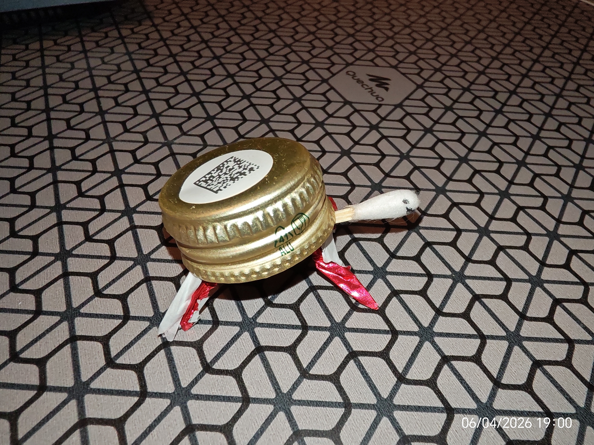 A close-up shot depicts a homemade turtle figurine made from a gold-colored metal bottle cap, a cotton swab, and red foil. The turtle is resting on a gray surface patterned with interconnected geometric shapes. The "Quechua" logo is visible in the upper right corner of the background. The date "06/04/2026 19:00" is stamped in the lower right corner. The location is Unknown, Unknown.