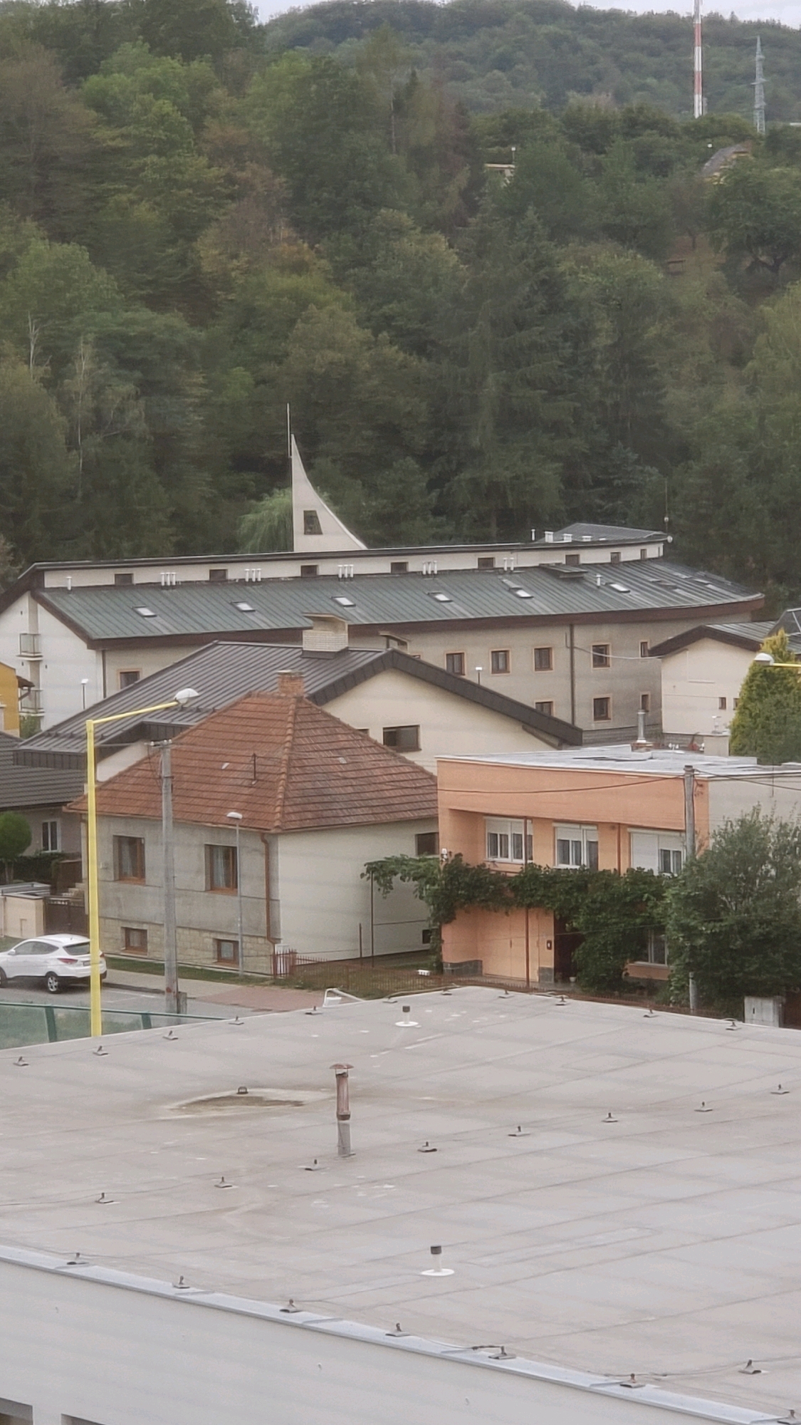 This is a slightly elevated, medium-shot, vertical, outdoor, daytime, full-color image of a residential area in Prešov, Slovakia, with a dense forest in the background. In the foreground, a large, flat, light gray rooftop with numerous small black anchors and a central chimney stack dominates the lower third of the frame. Beyond this rooftop, a row of buildings is visible, including a two-story house with a red-tiled, pyramid-shaped roof, a cream-colored exterior, and brown-trimmed windows. A yellow pole with a street lamp stands to the left of this house. Next to it is a larger, multi-story building with a dark gray, sloped roof featuring many dormer windows, and a distinctive white, pointed architectural feature rising from its roof. A smaller, tan-colored, two-story building with orange trim and white-framed windows is situated to the right of the red-roofed house. A white car is parked on a street to the left of the scene. The background is filled with a thick, green forest that covers a rolling hillside, with a tall, white and red radio tower visible at the top of the hill. The sky is overcast and pale.