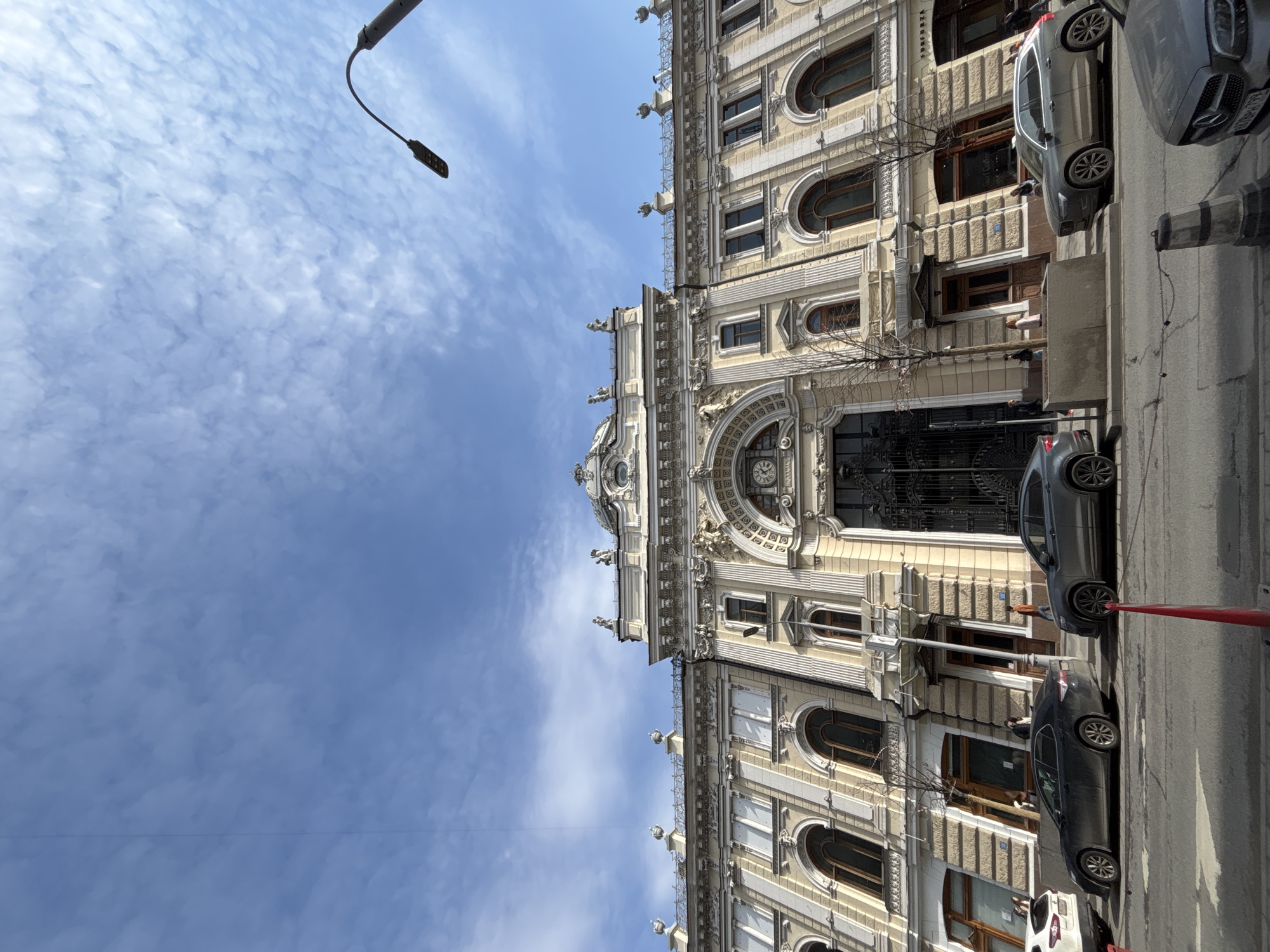 A street scene in Moscow, Russia features a grand, ornate building dominating the frame. The building, constructed from beige stone, displays intricate architectural details, including arched windows, decorative moldings, statues, and a clock. The building faces the street, where several cars are parked. Above, a bright blue sky is patterned with sparse clouds. A lamppost with a single lamp extends into the upper left corner of the frame.