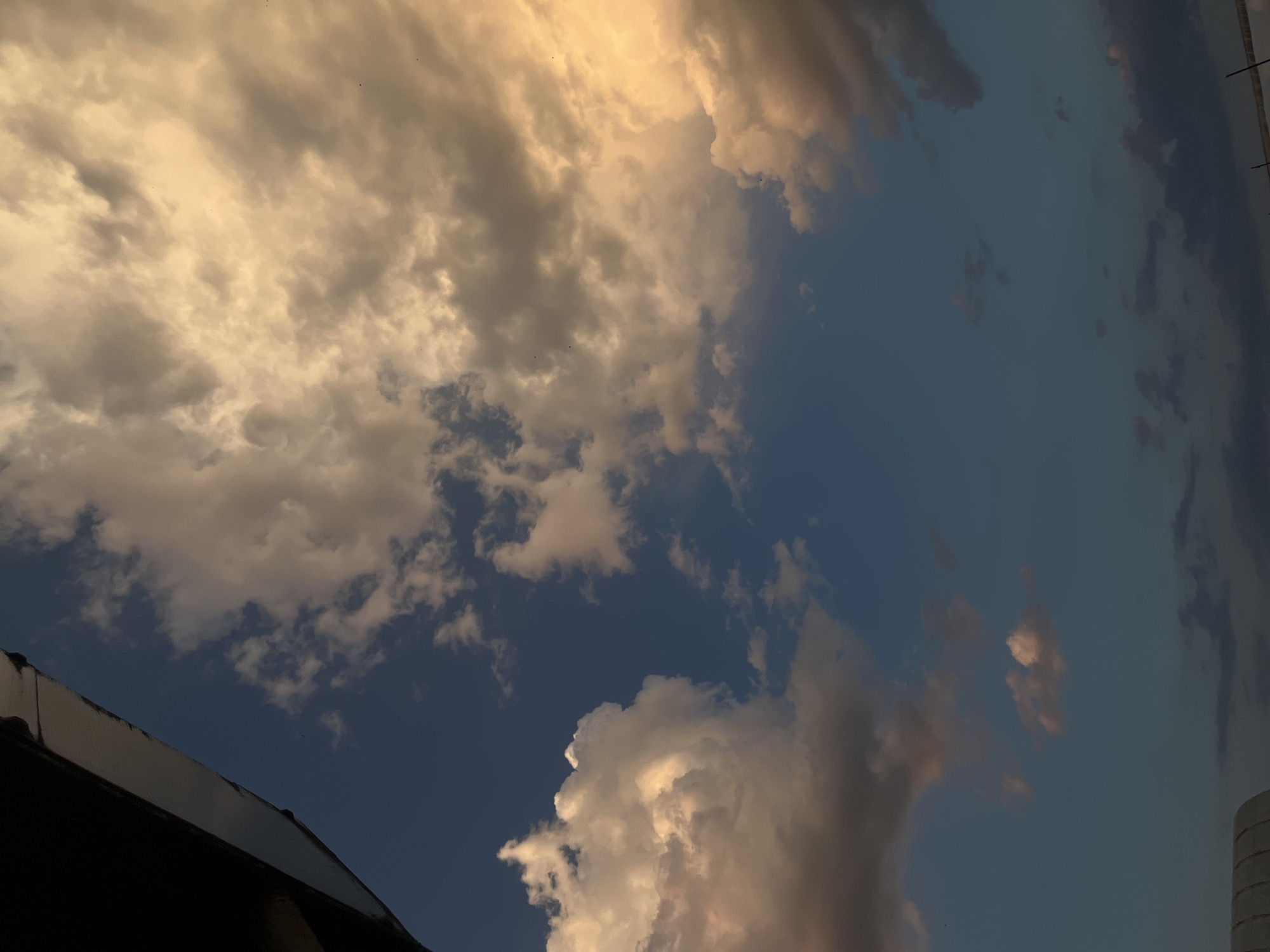 The image captures a section of the sky above what appears to be part of a building structure. The sky is partially covered with clouds, some illuminated with a golden hue, possibly from the sun. The blue sky is visible in between the cloud formations. Based on the prompt, this image is assumed to be taken in Novopokrovskaya, Russia. However, there are no visual elements in the image that independently confirm the exact location.