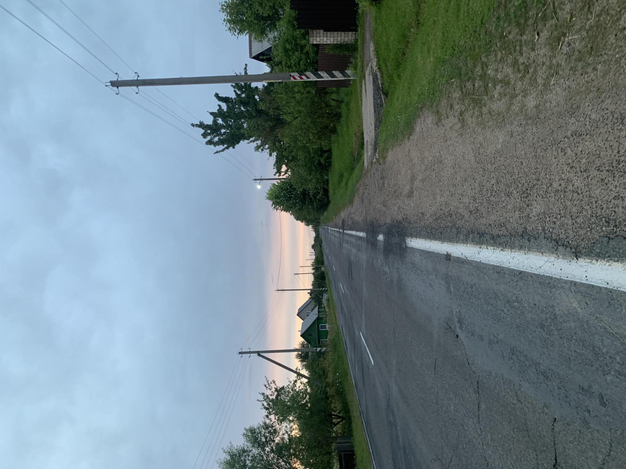 The image depicts a rural road in the Відзаўскі сельскі Савет, Belarus, at dusk. The road is paved and has white lane markings, with some cracks visible on the surface. To the left of the road, there are trees, green houses, and power lines. To the right, there is a gravel shoulder, grass, a fence made of brick and dark wood, and more trees. A power pole with striped markings stands on the right side of the road. The sky is overcast with a hint of pink and orange from the setting sun. The scene is quiet and appears to be a typical evening in a rural Belarusian village. There are no people or animals visible in the image.