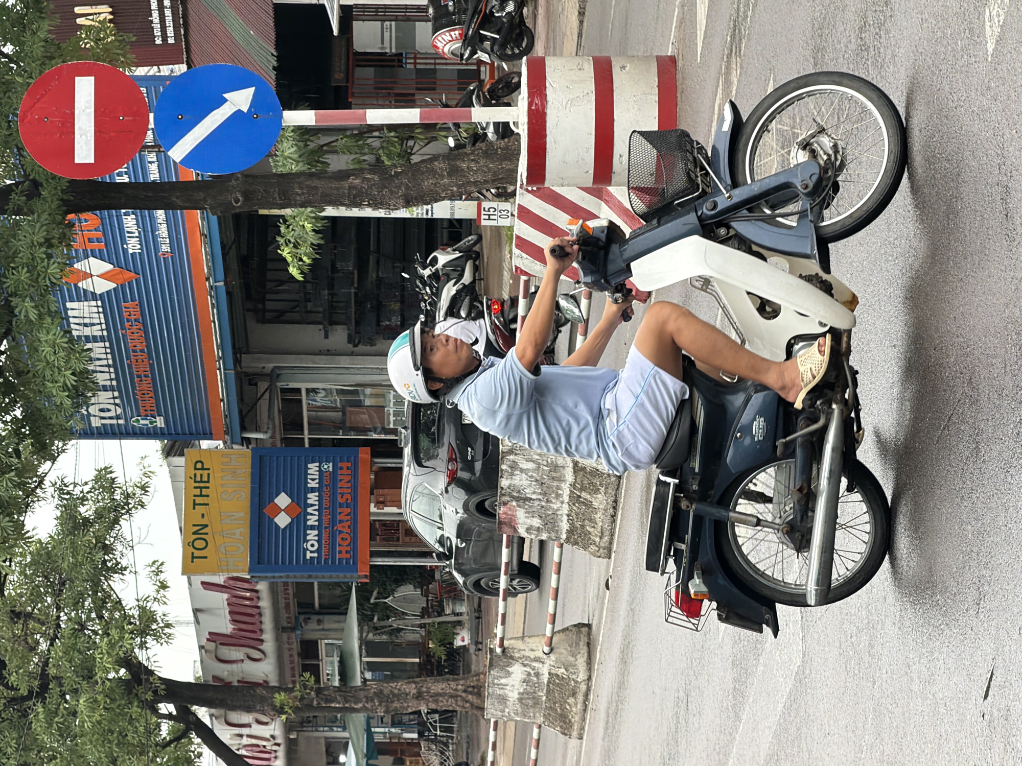 A man wearing a light blue polo shirt, white shorts, and a white and blue helmet rides a dark blue and white scooter. He is wearing tan sandals. The scooter has a black basket attached to the front and a red tail light. The man is looking to his left. The scooter is on a wet, grey asphalt road.
In the background, there are several buildings with signs. One sign in yellow and blue reads "TÔN NAM KIM" and "TÔN THÉP HOÀN SINH". Another sign above it is yellow and reads "TÔN - THÉP". There is also a large blue building with a sign that says "TÔN NAM KIM THƯƠNG HIỆU QUỐC GIA HOÀN SINH".
To the right of the buildings, there are two traffic signs on a pole. The top sign is red and round with a white horizontal bar, indicating "Do Not Enter". The bottom sign is blue and round with a white downward-pointing arrow, indicating "Turn Left".
A dark grey SUV is parked on the right side of the road behind concrete barriers. Several other scooters are parked in front of the buildings. The scene appears to be on a street in Nha Trang, Vietnam, indicated by the Vietnamese text on the signs. The sky is overcast and the road is wet, suggesting it may have recently rained.