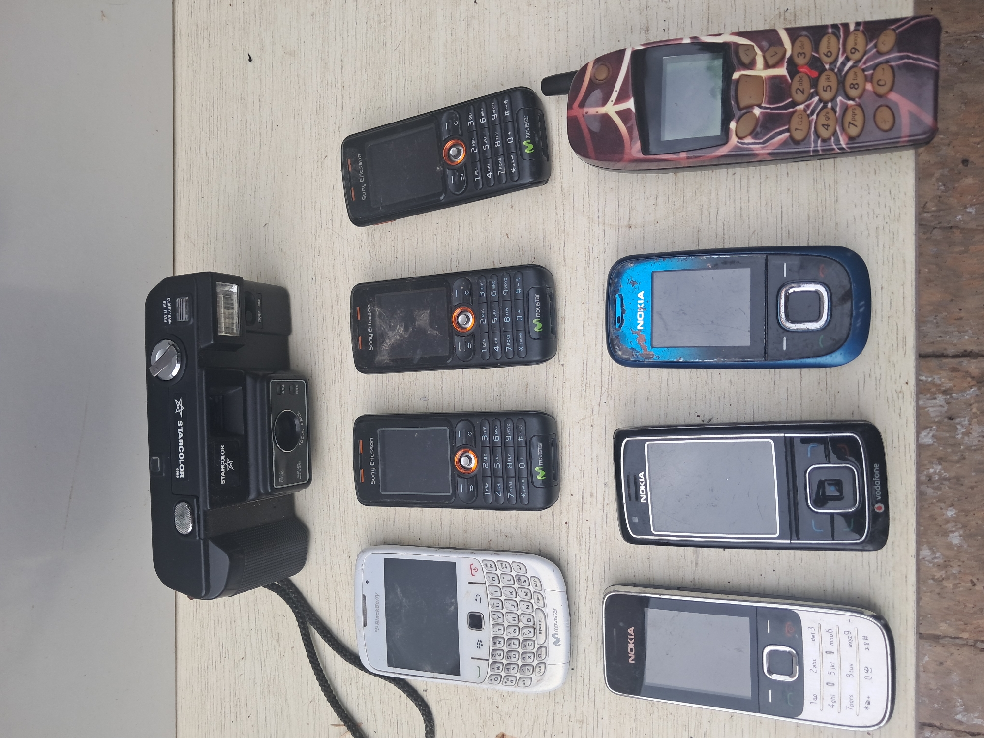 The image displays a collection of vintage electronic devices, including a black camera and several mobile phones, arranged on a wooden surface. The camera, positioned at the top, is a "STARCOLOR" model with visible buttons and a lens. Below the camera, a white Blackberry phone with a full QWERTY keyboard is situated to the left. To its right are three Sony Ericsson phones, each with a numeric keypad and orange accents. Further down, two Nokia phones are visible, one black and one blue. A distinctly patterned phone, with brown and red abstract designs, is placed to the right of the blue Nokia. The wooden surface appears weathered, suggesting an outdoor setting. The background is a plain, light-colored wall. No people are present in the image. The scene is set on a table, possibly outdoors, with natural lighting. The devices are dusty and appear to be old, indicating they may be part of a collection or for sale.