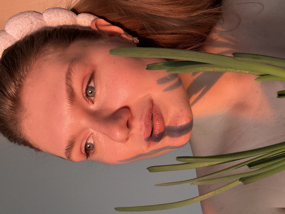 A woman with a pink headband and auburn hair is lying down. Her face is angled toward the camera, illuminated with a warm, rosy light. Her eyes are light, and her lips are tinged a slight purple. Shadows from long, green stalks lay across her face, adding an abstract dimension to the portrait.
