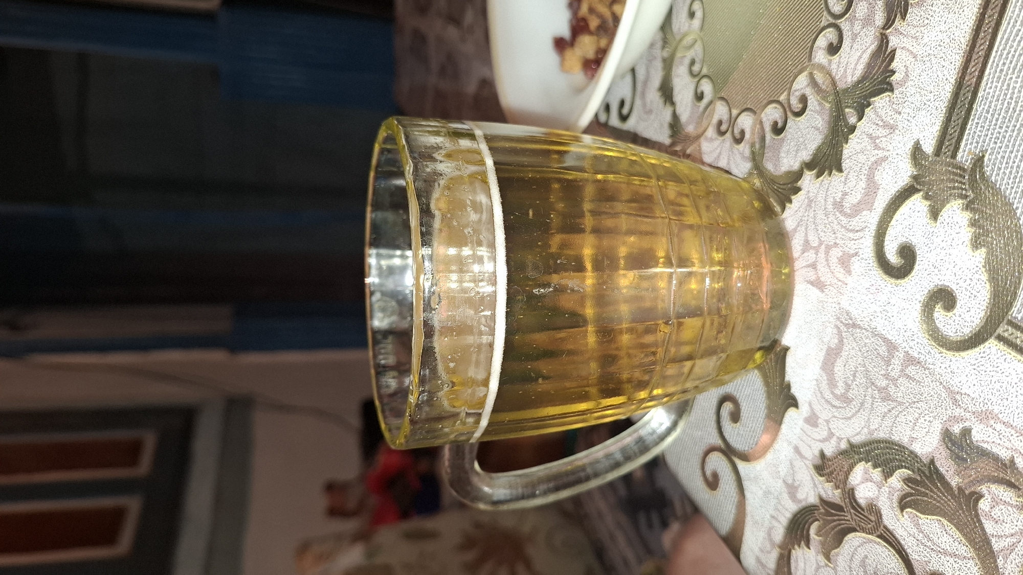 A close-up, eye-level shot captures a clear glass mug filled with a golden-yellow beverage, likely beer, with a thin layer of white foam at the top. The mug has a textured surface with horizontal and vertical ridges and a prominent handle on the left side. It rests on a patterned tablecloth with intricate swirls and floral motifs in shades of beige, white, and gold. To the right of the mug, a white bowl contains a mixture of small, irregular-shaped brown and red items, possibly nuts and dried fruit. The background is dark and out of focus, suggesting an indoor setting with muted lighting. The overall scene appears to be set in an informal environment. There is no indication of the specific location within Шұбарсу, Kazakhstan, beyond it being an indoor setting.