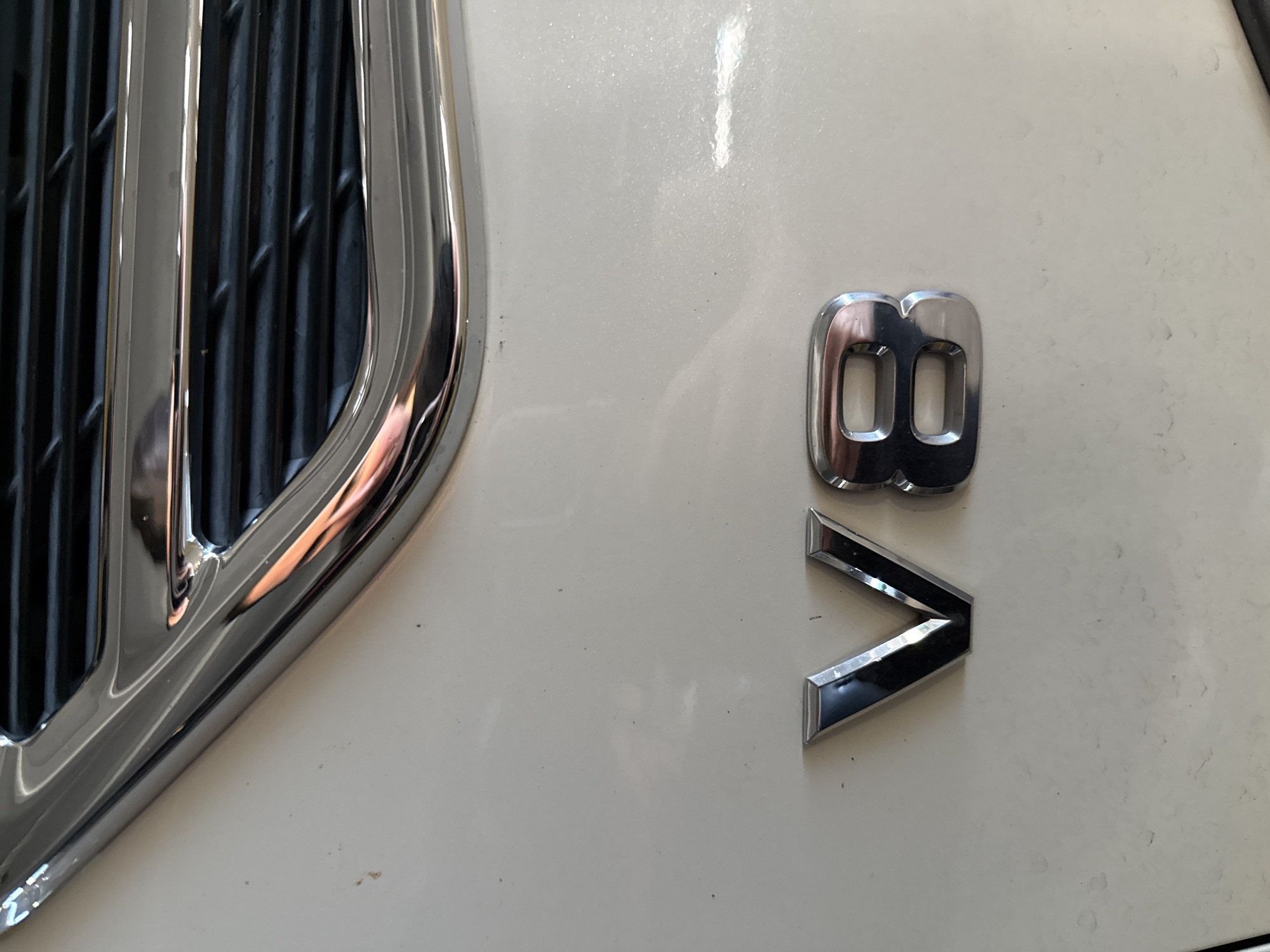 The image shows a close-up of the front of a vehicle, likely a car or SUV. The vehicle has a chrome grill with vertical bars visible on the left side of the frame. To the right of the grill, on the white exterior of the vehicle, are the letters "V8" in chrome. The "8" is positioned above the "V." The lighting appears to be bright and even, illuminating the chrome details and the white surface of the vehicle. There are no people visible in the image. The location, Hollywood Seminole Reservation, United States of America, is provided but not directly evident from the visual information in the photograph.