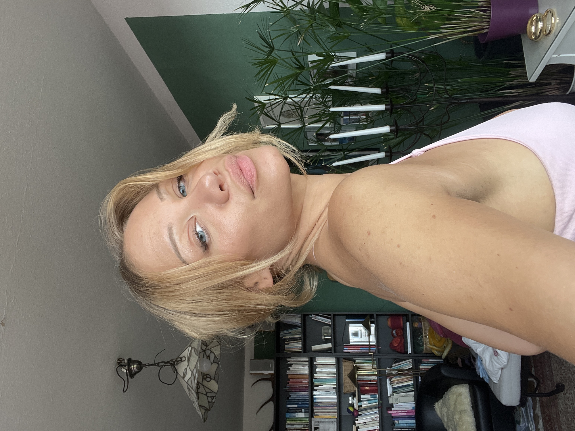 The media file is a vertical, eye-level, close-up selfie of a blonde woman with blue eyes. She is wearing a pink tank top. Her hair is blonde and falls across her face. Her lips are slightly parted and painted with pink lipstick. She has light skin with some freckles. The background is a room with a dark green wall and a bookshelf filled with books. There is also a plant with long, spiky leaves and a purple pot in the background. The lighting suggests it is daytime, and the overall mood appears relaxed and casual. There is no visible text in the image.