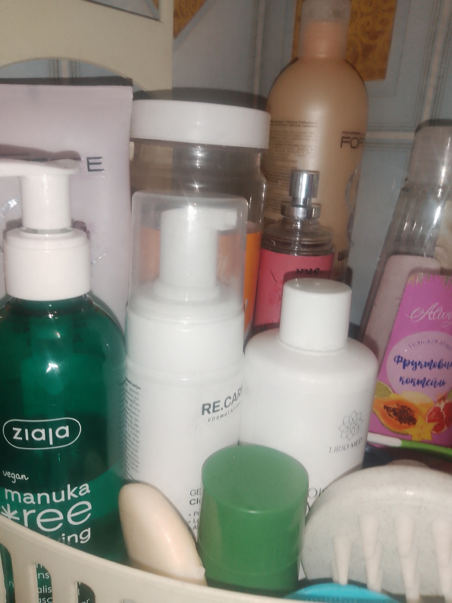 The image depicts a close-up, eye-level view of a collection of skincare and personal care products arranged in a basket or shelf. The lighting is somewhat dim and reflects off the glossy surfaces of the bottles and containers, creating highlights.
Prominently featured is a dark green bottle with a white pump dispenser, labeled "ziaja vegan manuka tree oiling." To its right, a white foaming dispenser with a clear plastic cap is visible, displaying the label "RE.CARE FORMULA." Next to this is a white bottle with a white cap, featuring a stylized lotus flower logo and the text "LIRIO MED."
Behind these items, several other products can be discerned. A large, pale orange bottle with a pump dispenser, possibly a lotion or shampoo, stands further back, with the word "FOR" visible on its label. To the right of this, a clear bottle with a silver pump, likely a perfume or body spray, is present. Further to the right, a pink package with stylized text and fruit imagery, possibly a soap or body wash, is visible, with "Aliv" and "Фруктові" (Fruits) legible. A small, white oval object, possibly a bar of soap or a cosmetic item, lies in front of the "RE.CARE" bottle. A bright green cap is positioned in the foreground, to the right of the "RE.CARE" bottle. At the very bottom, a white, textured object with multiple prongs, resembling a scalp massager, is visible. A blue object is partially visible underneath.
The background consists of a lightly striped wall or surface and what appears to be the edge of a shelf or cabinet. The image does not contain any explicit indicators of its location within the city of Chernivtsi, Ukraine, nor does it show any people or interactions. The scene is static and focuses solely on the arrangement of the consumer products.