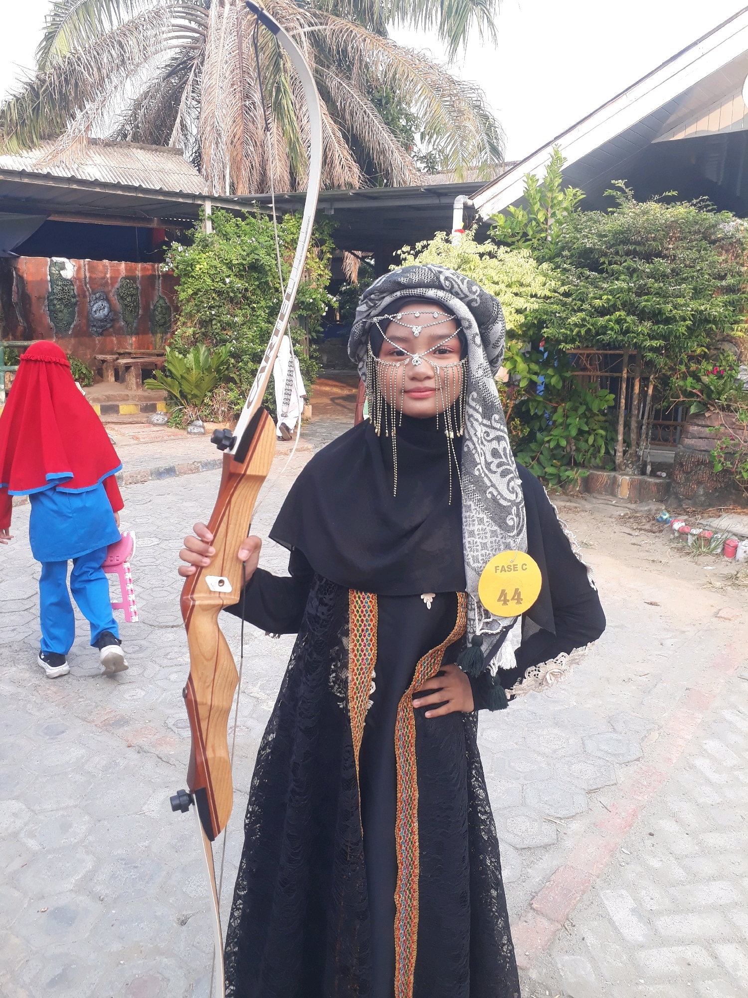 A young girl, identified by a yellow circular tag on her chest displaying "FASE C" and the number "44", stands outdoors in Pangkalpinang, Indonesia, holding a wooden recurve bow. She is wearing a black abaya adorned with a colorful trim along the front opening and cuffs, and a patterned headscarf. Her face is decorated with a metallic headdress featuring intricate beadwork and dangling chains. To her left and slightly behind her, another child is visible from the back, wearing a red head covering, a blue top, and blue pants, and white athletic shoes. The background shows a palm tree with dry fronds, a structure with a corrugated metal roof, and lush green vegetation. The ground appears to be paved with interlocking concrete blocks and some dirt. The overall scene suggests an outdoor event or competition taking place in a tropical or subtropical environment.