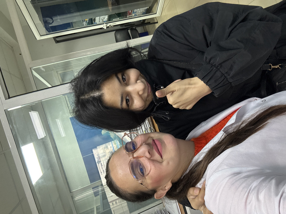 This image depicts two individuals, appearing as women, engaged in a selfie within an indoor office setting in Meknes, Morocco. The person on the left wears a white lab coat over an orange textured sweater and light pink hexagonal-framed glasses, with her dark hair pulled back. She smiles and has her right arm extended around the shoulder of the person on the right. The individual on the right, with dark, shoulder-length hair, is dressed in a black puffer jacket, smiles, and gives a "thumbs up" gesture with her right hand. The background features modern glass partitions framed in white, revealing a stack of documents on a desk, an office chair, and a blue-toned image or exterior view through what appears to be a window showing distant urban buildings. The specific location or address within Meknes cannot be identified from the visual information provided.