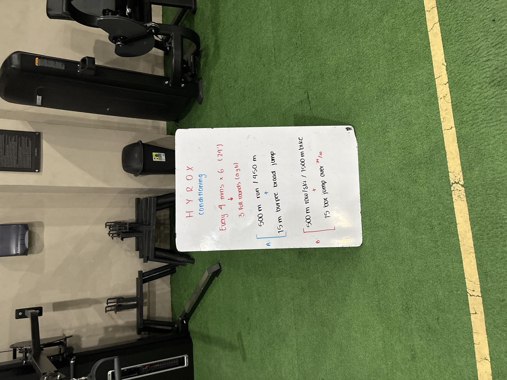 This media file captures an indoor gym setting, likely in San Bernardino Tlaxcalancingo, Mexico, with a focus on a whiteboard displaying a HYROX conditioning workout. The scene features green artificial turf flooring with a yellow line, and various pieces of fitness equipment including what appears to be a rowing machine or ski erg, and potentially a stationary bike or treadmill in the background. A black bin is also visible on the floor.
The main subject is a white whiteboard with workout instructions written in black and red marker. The text outlines a "HYROX conditioning" session structured as "Every 4 mins x 6 (24')" for "3 full rounds (a&b)". The workout is divided into two parts, A and B.
Part A includes:
- "500m run / 450 m"
- "+"
- "15m burpee broad jump"
Part B includes:
- "500 m row/ski / 1500m bike"
- "+"
- "15 box jump over" with "24/20" likely indicating heights in inches for the box jumps.
The word "ONSIDE" is visible on the side of one of the black fitness machines. The image is taken from a slightly elevated perspective, looking down at the whiteboard and the gym floor. No people are present, and the time of day, weather, or emotions are not discernible.