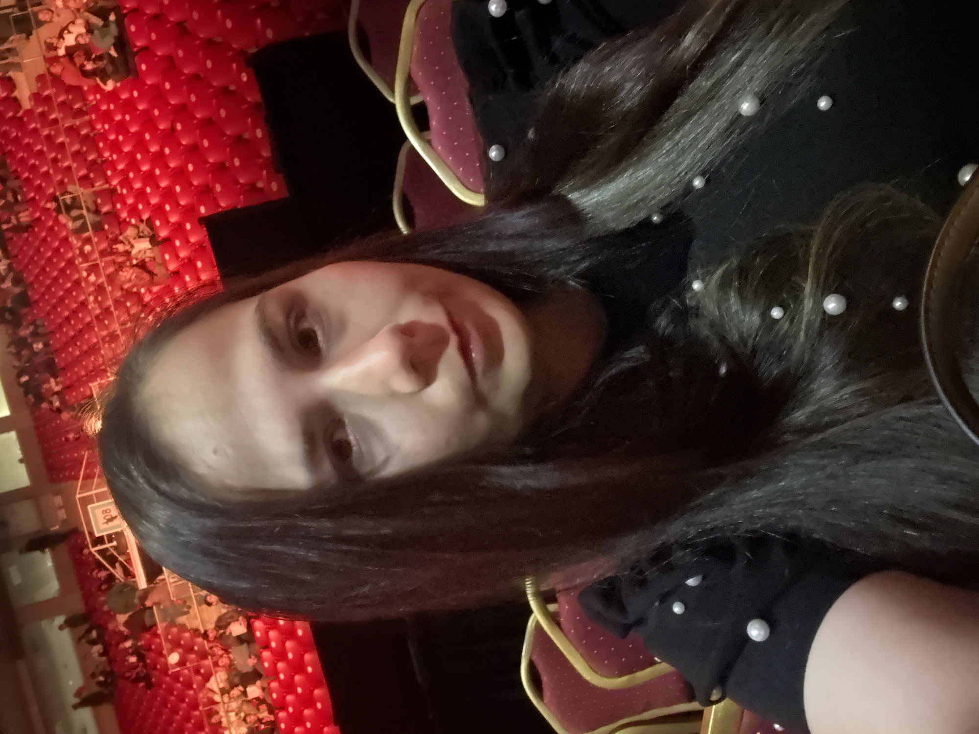 The image is a rotated, close-up selfie of a young person, likely female, captured indoors within a large auditorium or event hall in Sofia, Bulgaria. The subject is positioned in the foreground, looking directly at the camera with a neutral to slightly smiling expression. She has long, dark hair, partially adorned with small, white pearl-like beads. She is wearing a dark-colored top, which also features similar white pearl embellishments on the visible sleeve.
The background displays numerous rows of bright red seats, typical of a theater, concert hall, or conference venue. Many of these seats have white dots, which could be a design feature or reflections. Yellow structural frames are also visible on some seats. Faint outlines of other people are discernible in the distant audience areas. The lighting suggests an indoor setting, likely during an event. A sign with the number "408" is faintly visible in the background, possibly indicating a row or section within the venue. The original image is rotated 90 degrees clockwise.