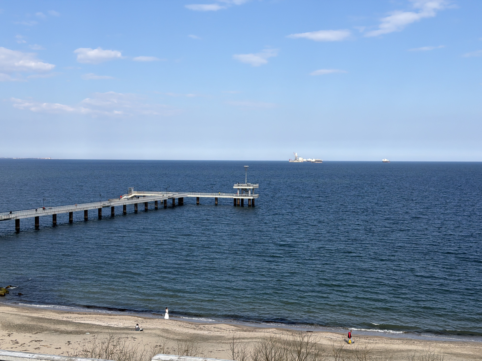 The image depicts a coastal scene in Burgas, Bulgaria. A long, concrete pier stretches into the sea from the beach, ending in a small platform structure. People can be seen walking on the pier. In the distance, several ships are visible on the horizon. On the sandy beach, several individuals are present; one person is wearing a white dress. Some bare trees are in the foreground. The sky is blue with scattered clouds. The sea is moderately choppy.