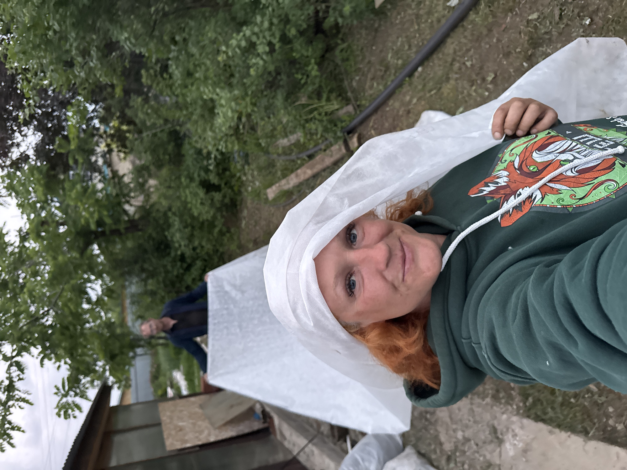 In an outdoor scene that appears to be a yard or garden area, a woman with red hair is taking a selfie. She is wearing a green hooded sweatshirt with a design featuring a fox and is partially covered by a white material, possibly a tarp or protective covering, which is also draped over her head like a veil. In the background, a man in a dark jacket is holding up another section of the white material, creating a tent-like structure. The environment is somewhat cluttered, with a small building or shed visible to the left, wooden boards, and lush green foliage surrounding the area. The ground is a mixture of dirt and grass. The overall impression is of a candid moment during some outdoor activity or project. The location is specified as Chaikovsky, Russia.