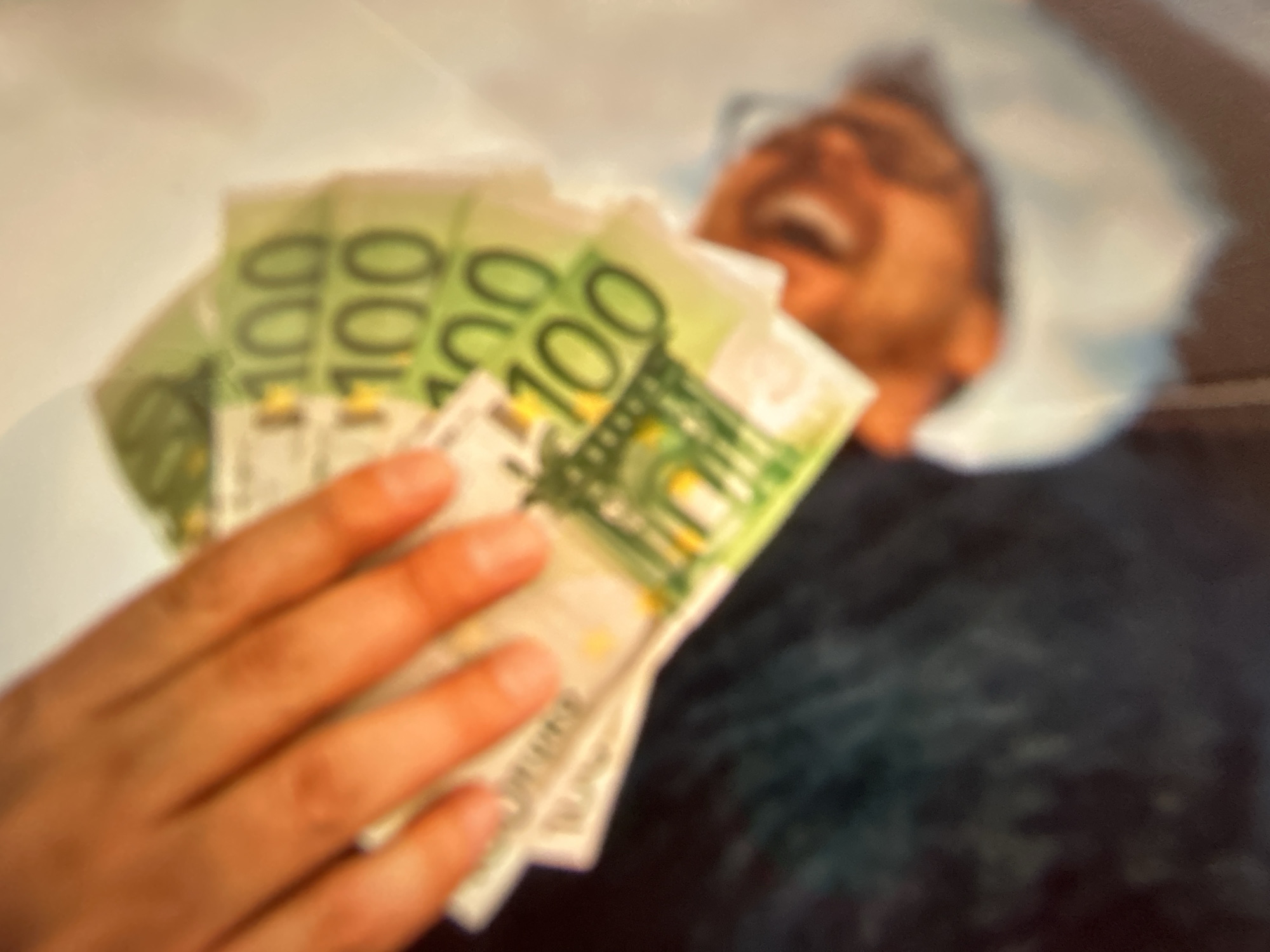 A person's hand is fanned out, holding a stack of Euro banknotes. The banknotes appear to be 100 Euro notes, indicated by the visible "100" denomination printed on them. The image is blurry, but suggests the notes are green and white. A portion of a face is visible beneath the banknotes, with a blurred orange tone. The scene is set against a dark background with some lighter, out-of-focus elements. There is no information in the image to suggest a location in Gelendzhik, Russia.