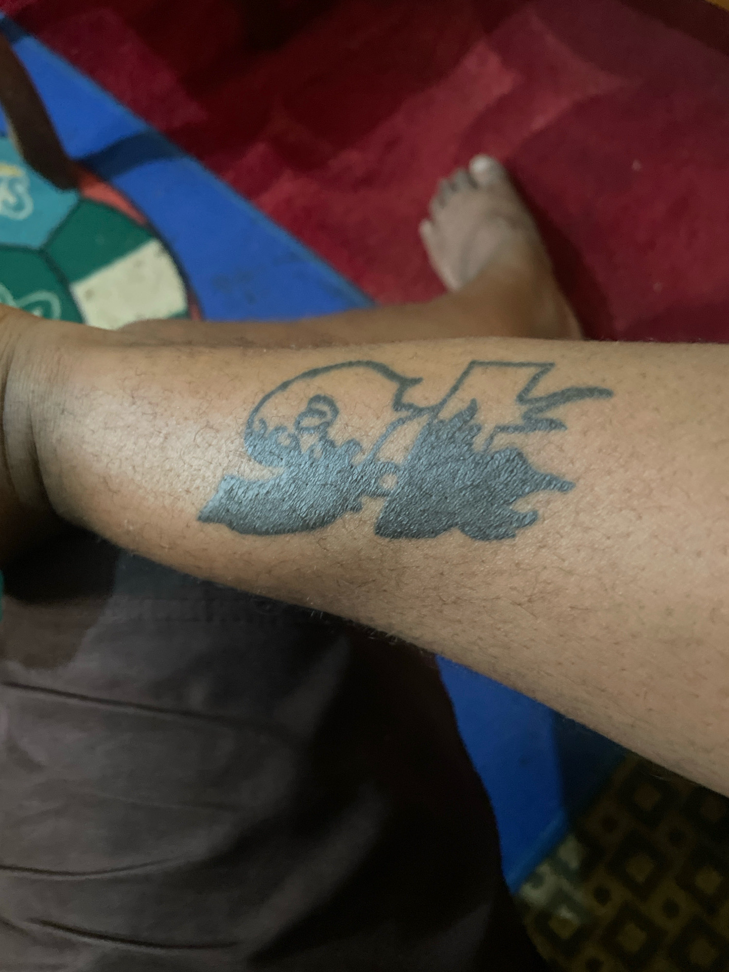 A tattoo in black ink is visible on a person's forearm. The tattoo appears to be a stylized depiction of two figures or possibly animals in profile facing each other, with jagged lines extending from the area between them, suggesting an energetic or combative interaction. The skin around the tattoo is lightly tanned and shows some fine hairs. The background consists of a red, textured surface, possibly a rug or blanket, and a sliver of a blue surface. The person's foot, with visible toes, is also partially visible in the upper right quadrant, resting on the red surface. A darker, draped fabric is visible in the lower left of the image. No specific location within Jos, Nigeria, can be identified from the provided visual information.