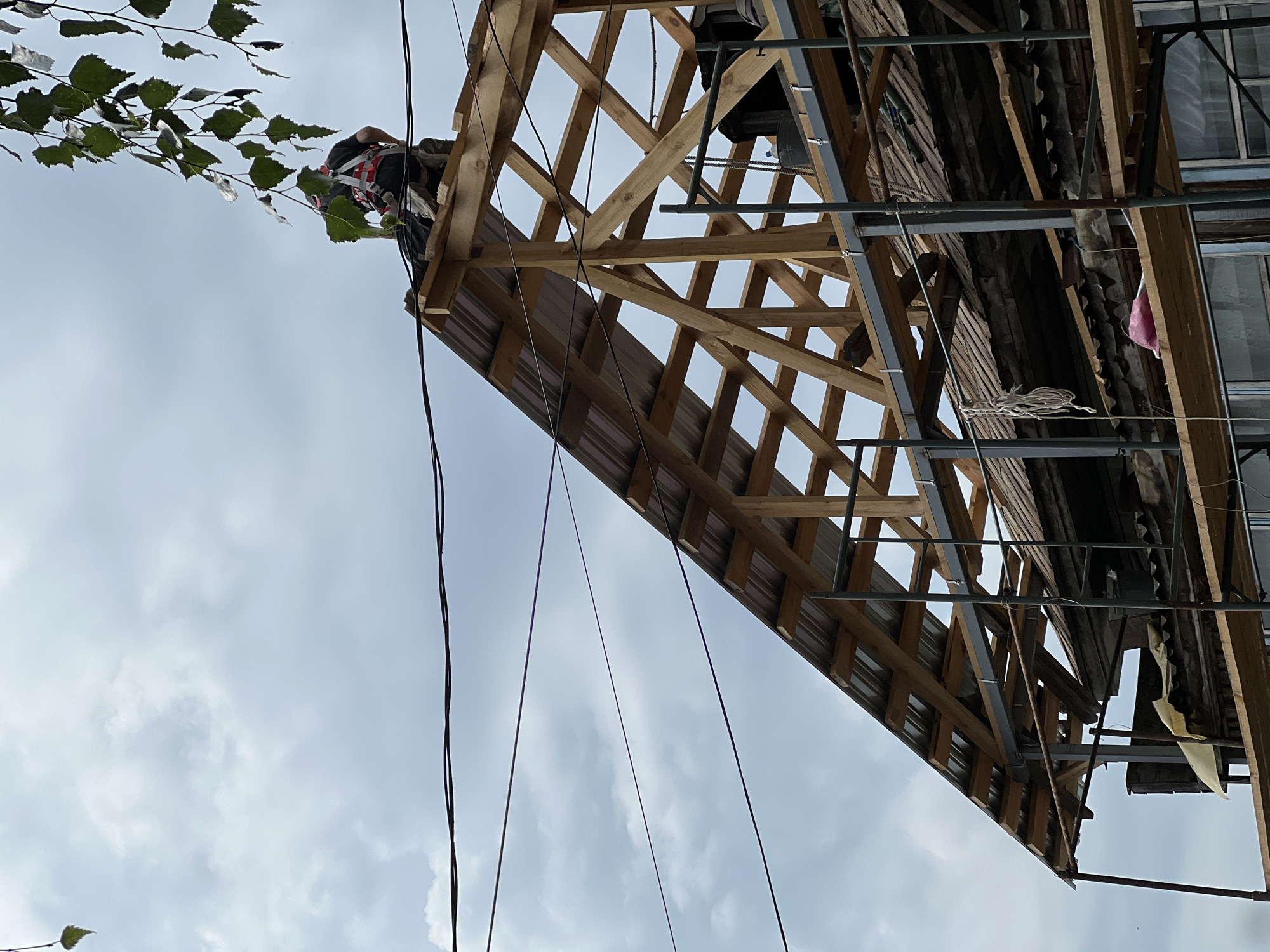 The image depicts a construction or renovation scene focused on the roof of a building in Biysk, Russia. A construction worker, wearing a safety harness, is positioned on the roof, which is in a state of partial construction. The roof structure is made of visible wooden beams and what appears to be corrugated metal sheeting. Scaffolding surrounds a portion of the building, with wooden planks and metal supports visible. Power lines stretch across the sky in the background, and a few branches from a tree hang into the frame from the upper right corner. The sky is overcast with visible clouds. The overall impression is one of ongoing construction work on an older building.