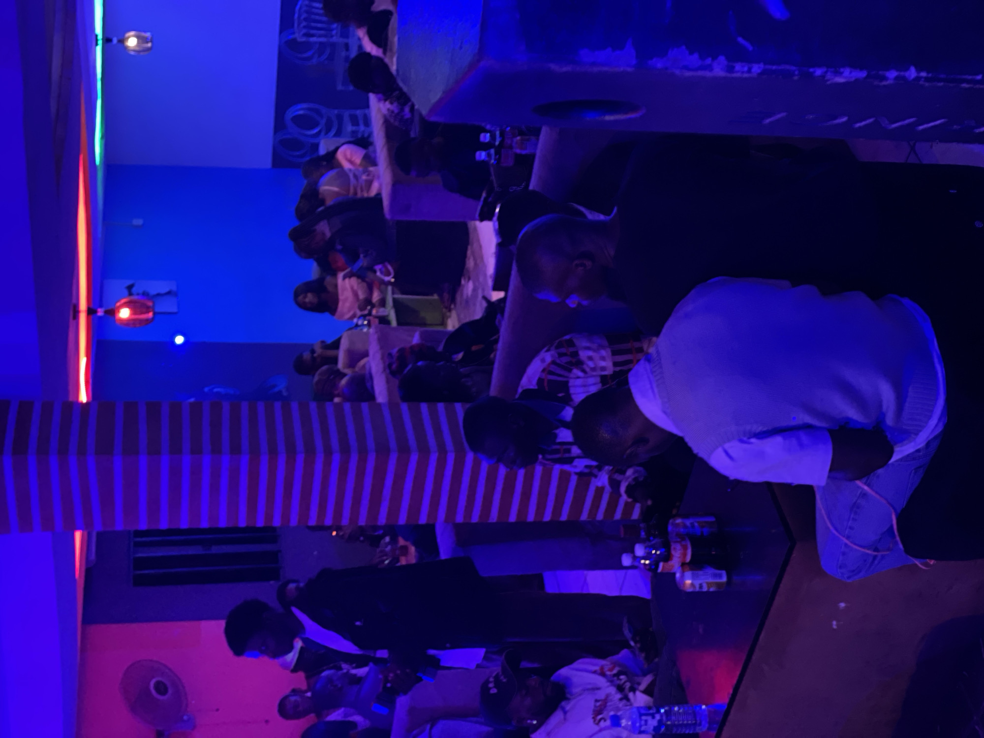 The image depicts an indoor setting, likely a nightclub or lounge, illuminated by vibrant blue and purple lights. In the foreground, a person in a white vest and blue jeans is seated facing away from the camera. To the left, a black table holds several bottles and cans of beverages, including a bottle of water. A person wearing a black jacket and white shirt is standing to the left of the table, partially visible. In the background, several other individuals are seated and standing, some engaged in conversation. The environment features plush seating, a striped column, and decorative lighting. The overall atmosphere suggests a social gathering or nightlife scene in Durumi, Nigeria.