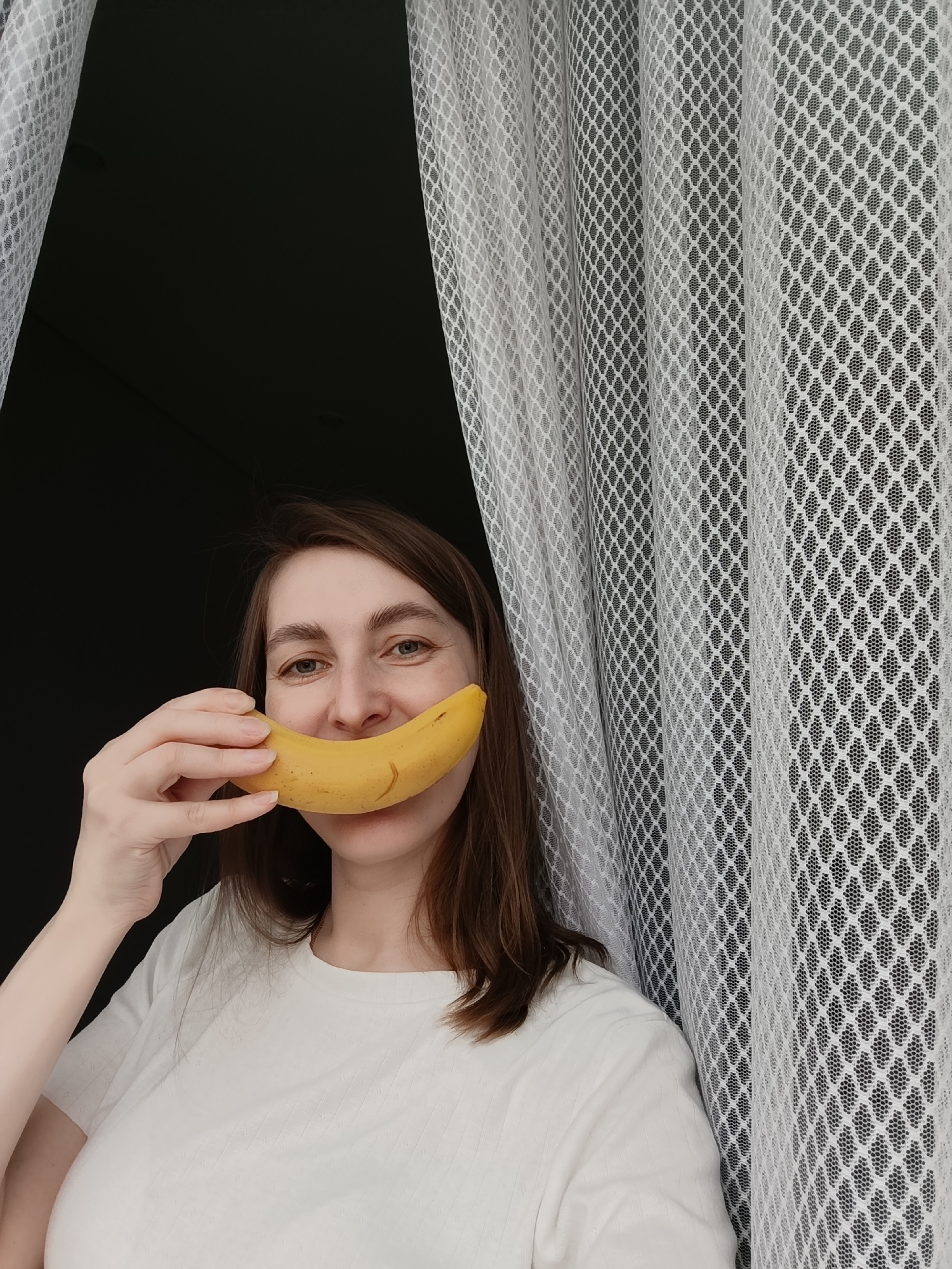 A woman stands indoors in front of a white lace curtain, holding a yellow banana in front of her mouth to give the appearance of a wide smile. The background beyond the curtain appears to be a dark or dimly lit room. The woman is wearing a white t-shirt. The location may be Chaikovsky, Russia, but the provided image does not show enough detail to confirm it.