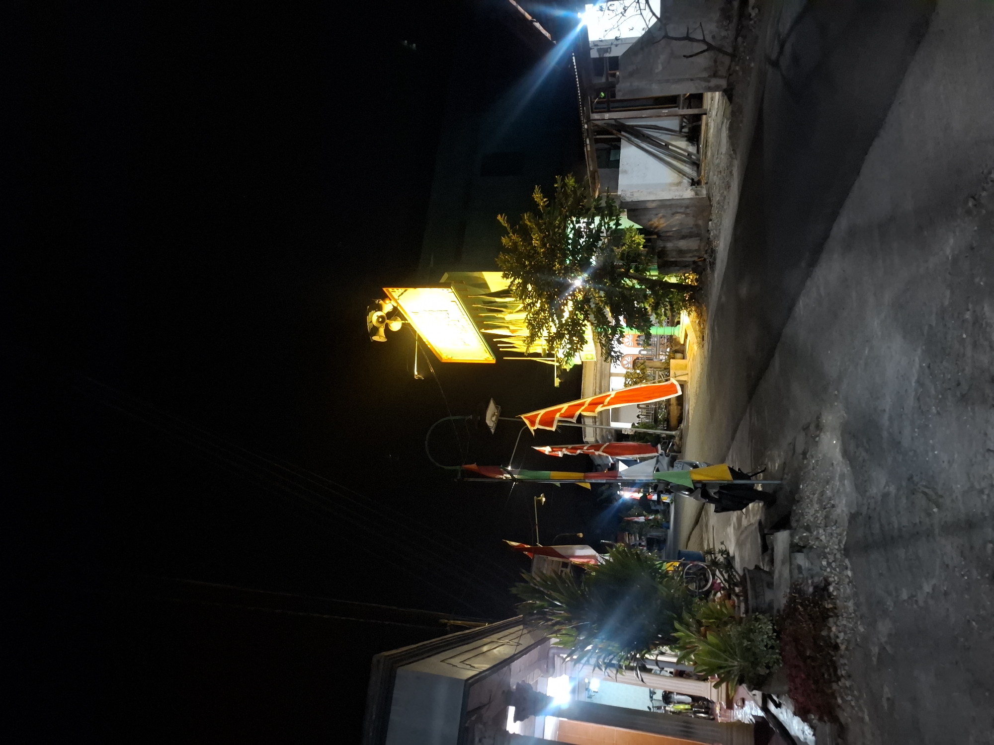 The image depicts a street scene at night in Bulubrangsi, Indonesia. The foreground shows a paved road with a slight incline. To the left, a building with a gray, textured facade is visible, illuminated by warm light emanating from within. In front of this building, there are potted plants and a small pile of gravel. Several flags, predominantly red and white, are mounted on poles along the side of the road.
Further down the road, a motorcycle is parked. On the right side of the image, another building is partially visible with a light-colored facade and some wooden structures against it. A large, bright yellow sign with text is mounted on the facade of a building in the background, illuminated by lights from above. A tree with green foliage is situated in front of this sign, also lit by artificial light. Electrical wires are visible overhead, spanning across the dark night sky. The overall atmosphere is of a quiet street at night, likely in a residential or commercial area given the building structures and signage.