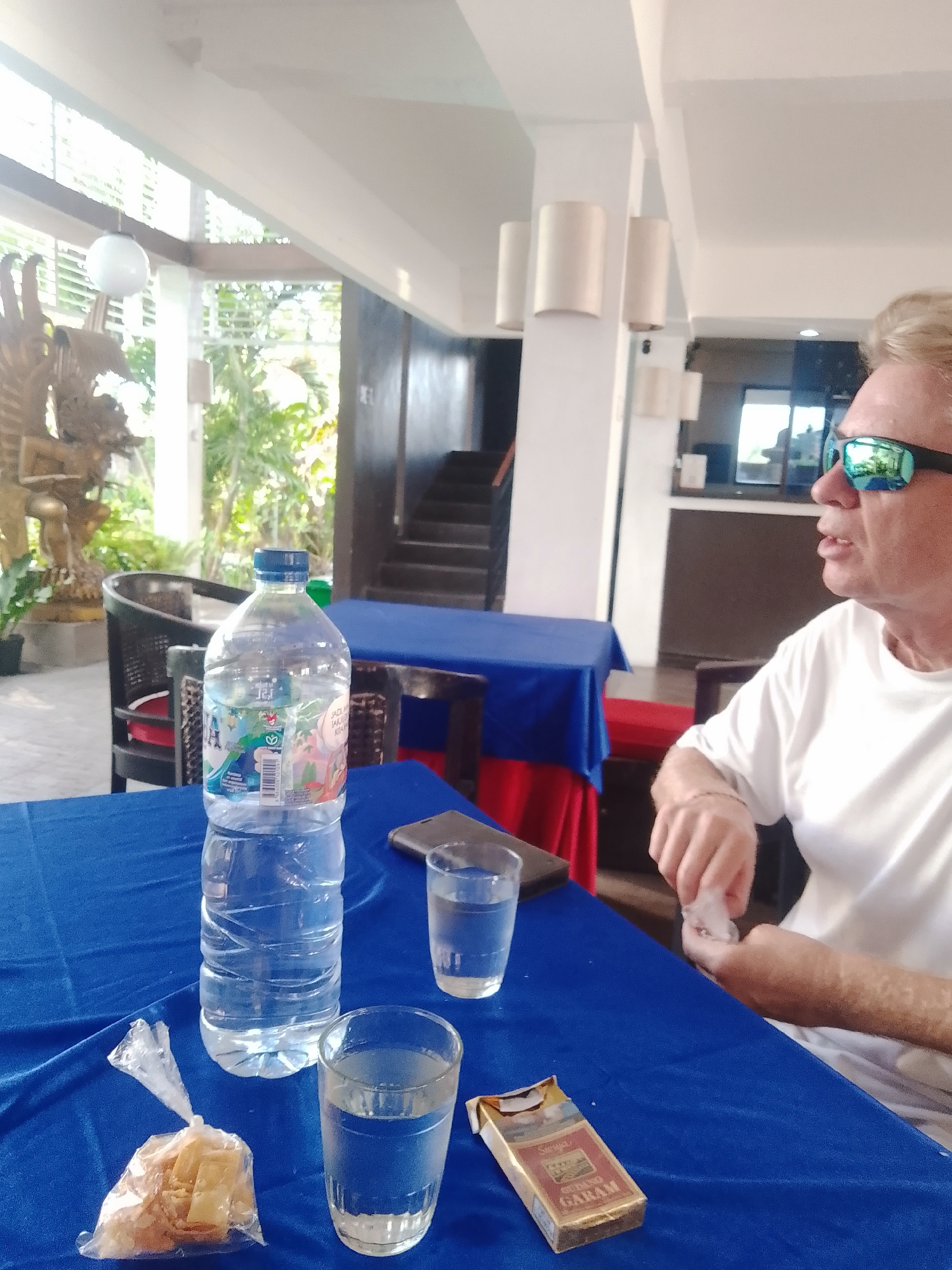 A man with blond hair and sunglasses sits at a blue-covered table in an open-air restaurant or cafe in Denpasar, Indonesia. He is wearing a white t-shirt and appears to be holding a cigarette. On the table in front of him are two glasses of water, a partially opened pack of Surya Garam cigarettes, a small plastic bag containing some snacks, and a bottle of Aqua water. In the background, there are tables draped with blue and red cloths, chairs, and a staircase leading to an upper level. Lush greenery is visible through large windows, suggesting an outdoor or semi-outdoor setting. The overall atmosphere appears relaxed and casual.