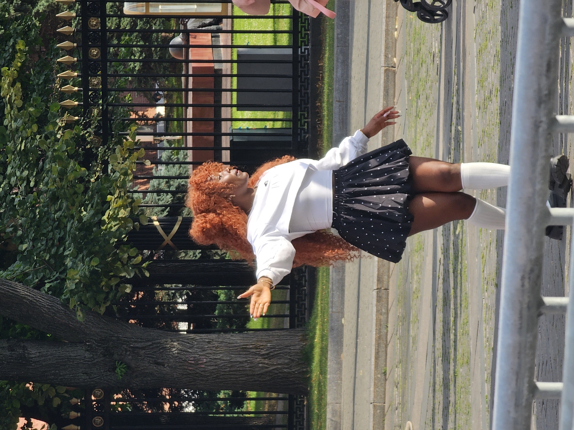 A Black woman with bright orange, voluminous curly hair and a white long-sleeved cropped top, black polka-dot pleated skirt, and white knee-high socks stands on a paved area in a city. Her right arm is extended outwards and slightly upwards, with her palm open. She is facing left, with her body angled to the right. Behind her is a black metal fence with ornate gold finials. Beyond the fence, trees, grass, and some red brick structures are visible. A stroller with large wheels is positioned to the far right of the frame. The ground is paved with cobblestones and has patches of grass growing through. The image appears to be taken during the daytime, with sunlight casting shadows. The presence of the fence with decorative elements and the surrounding architecture suggest the location could be a park or historical site within Moscow, Russia.