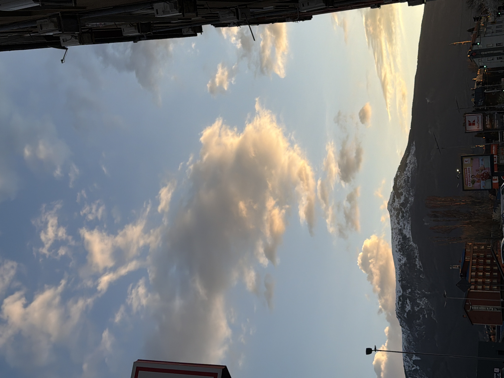 The image shows an upward view of the sky in Sofia, Bulgaria, with clouds illuminated by the setting sun. A mountain range is visible in the distance. Buildings and advertising billboards line the right side of the frame. From the lower-left to the top-left, there are additional buildings.