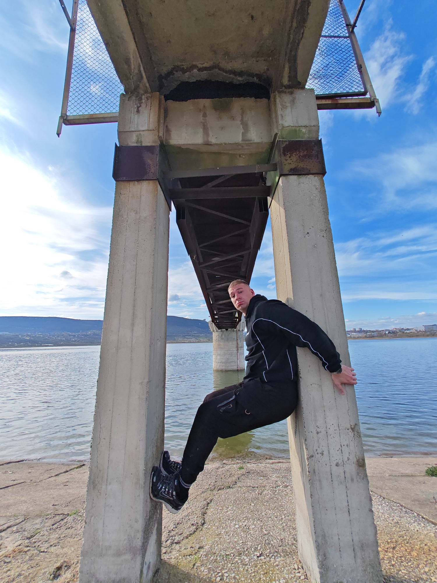 A man in black athletic wear is sitting sideways between two concrete pillars of a bridge. The bridge spans a body of water with hills in the background. The sky is blue with scattered clouds. The man is leaning against the right pillar with his feet braced against the left pillar, holding himself in place. The location is likely the bridge over Lake Shumen in the city of Shumen, Bulgaria.