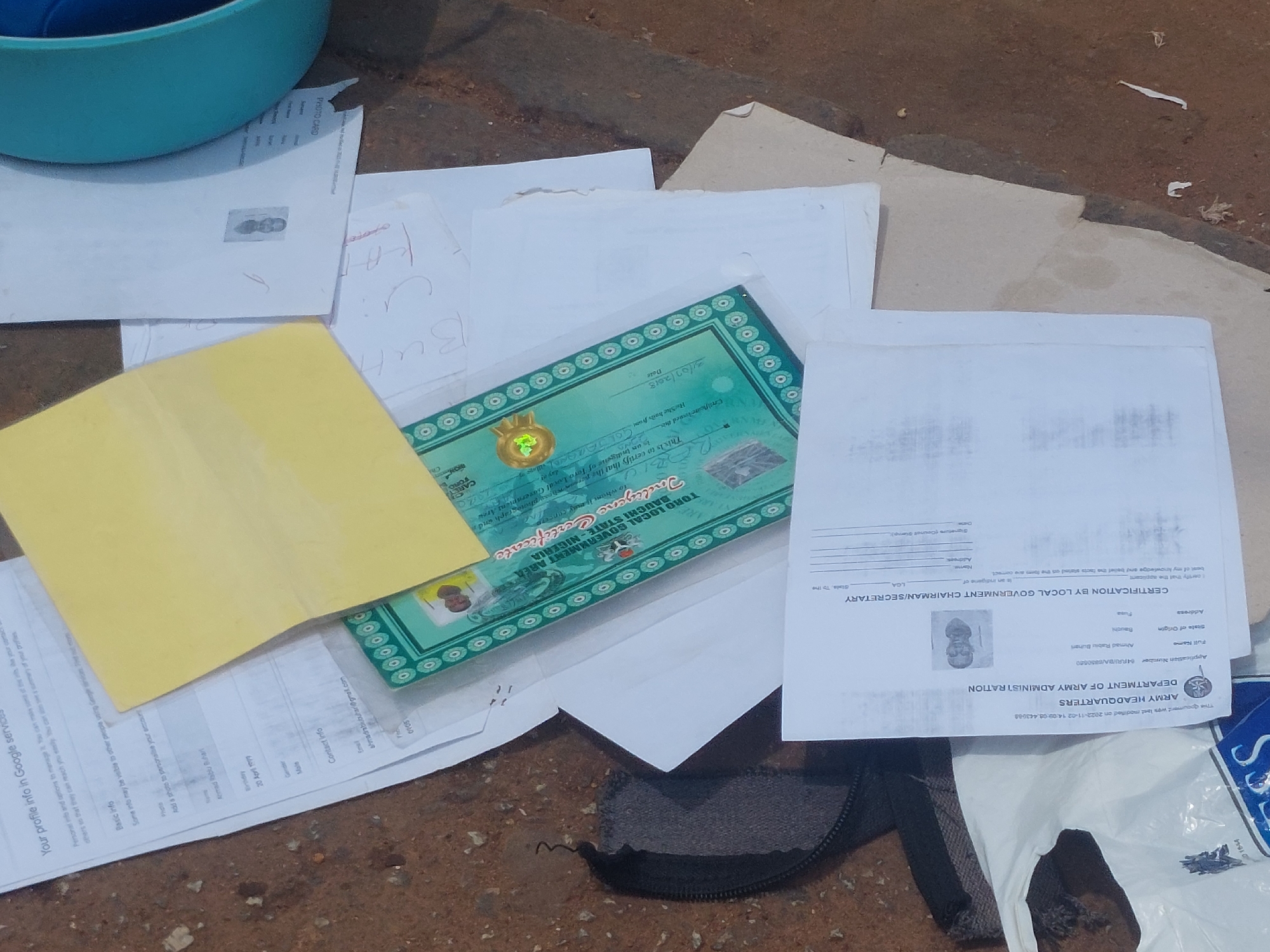 The image is an overhead shot of several documents scattered on a rough, outdoor surface that appears to be a mixture of dirt and pavement. In the upper left corner, a portion of a turquoise plastic bowl or container is visible. The main focus is on a collection of papers, some of which are official documents.
One prominent document is a green certificate with decorative borders and seals, labeled "TORO LOCAL GOVERNMENT AREA, KWARA STATE, NIGERIA, INDIGENE CERTIFICATE." It contains fields for "Date," "Full Name," and other information, with a picture of a person at the bottom. Another document, a white paper with black text, is titled "CERTIFICATION BY LOCAL GOVERNMENT CHAIRMAN/SECRETARY" and also includes fields for "Full Name," "Address," "LGA," and a photograph. Other papers include what looks like a yellow envelope, plain white sheets with handwritten notes, and a paper with a black and white photo of a person. The scene is disorganized, with papers overlapping and some appearing creased or torn, suggesting they have been handled and are not neatly stored. The ground surface is textured and appears to be outdoors.