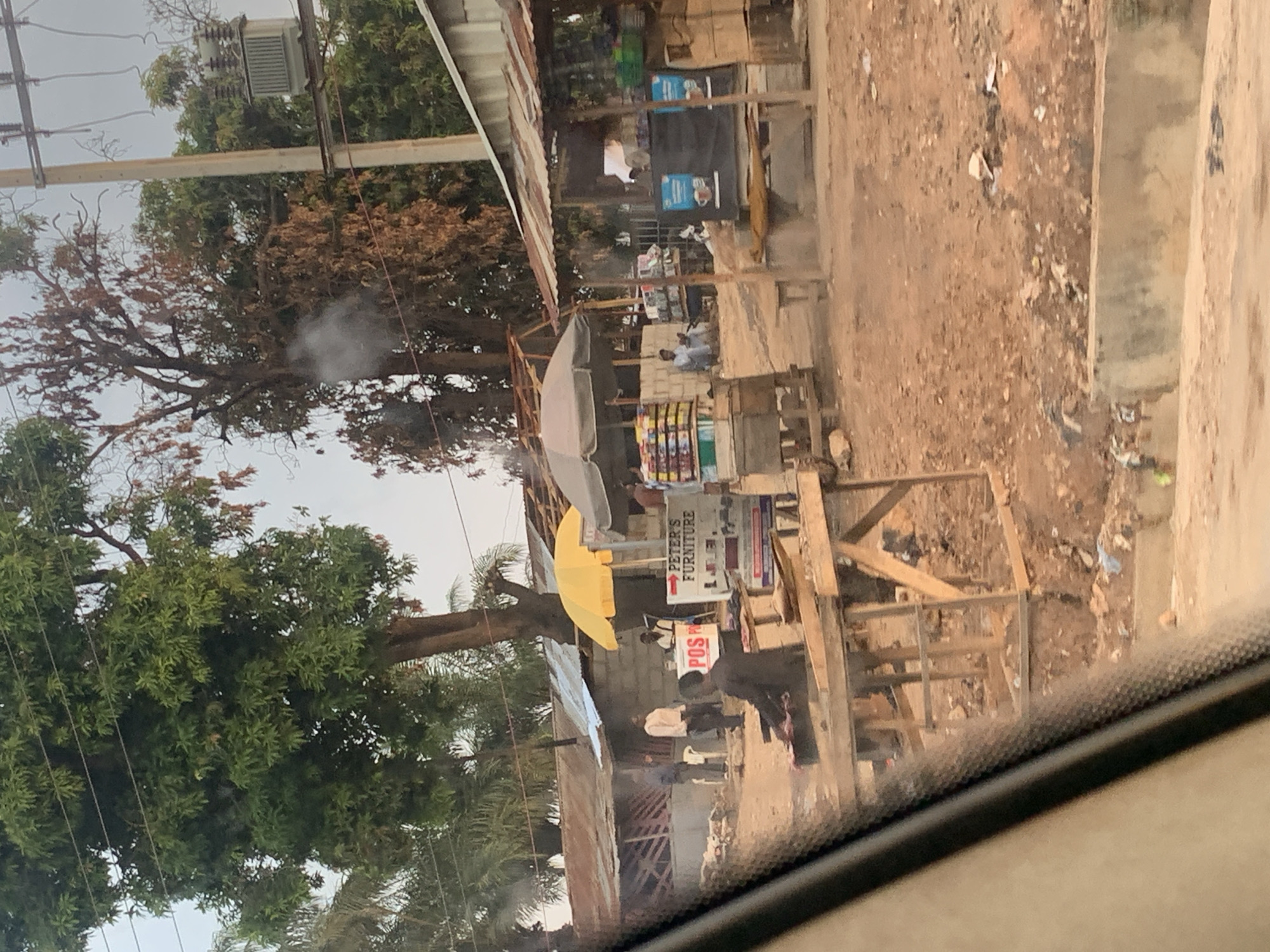 The image shows a street scene in Keffi, Nigeria. There are several wooden structures, possibly market stalls, lining the street. One stall has a sign reading "Peter's Furniture" with an arrow pointing towards it. A yellow umbrella provides shade for another stall. People are standing and walking near the stalls. The street appears dusty and unpaved. Trees are visible along the side of the road, with power lines overhead. The image was likely taken from inside a vehicle.