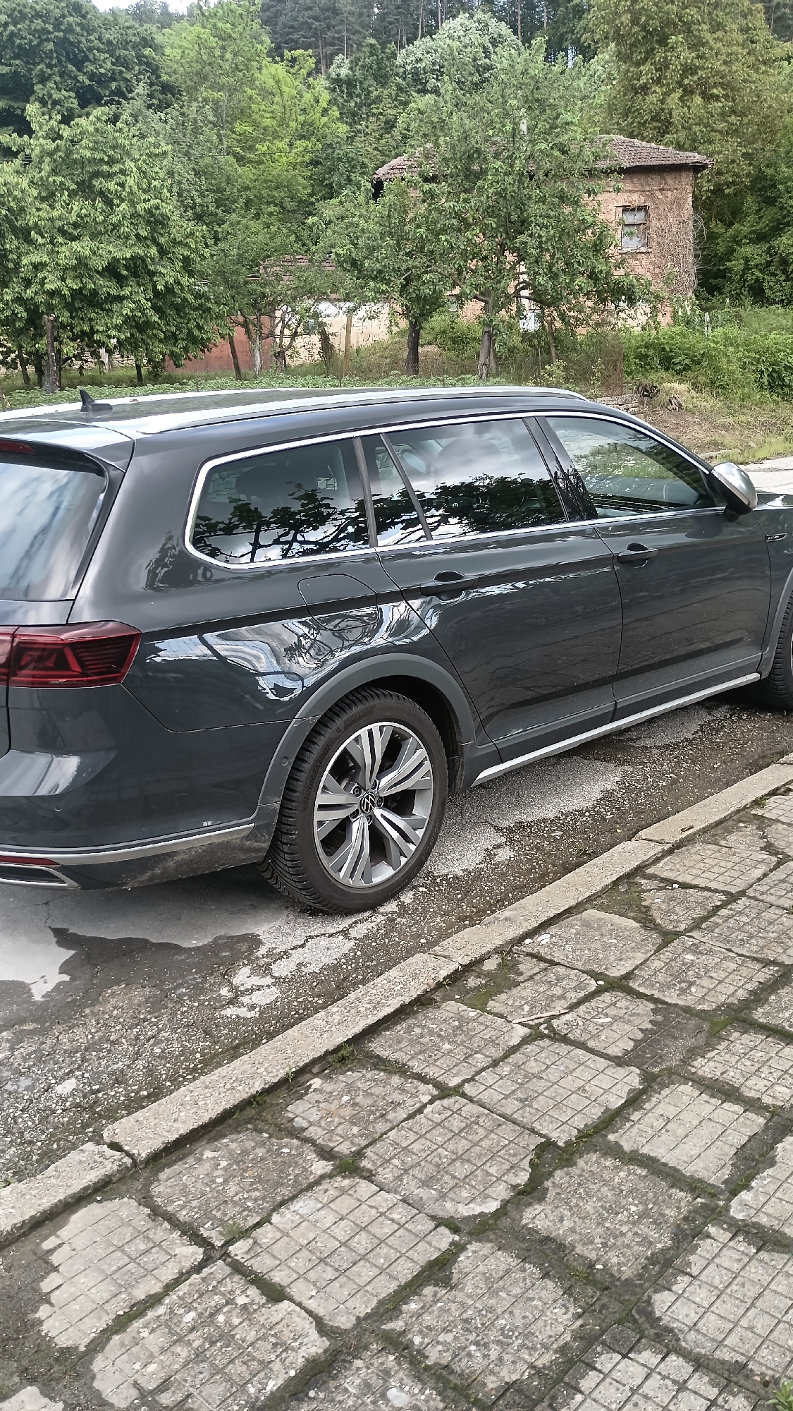 The image shows a gray Volkswagen Passat Alltrack parked on a wet street in Muzga, Bulgaria. The car is facing right, and a sidewalk made of square, concrete blocks is to the right of the car. Behind the car are trees and a brick building with a window. The sky is overcast.