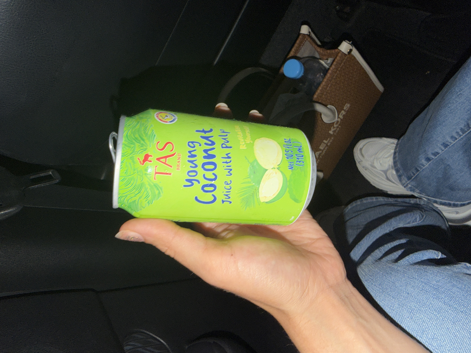 A hand holds a green aluminum can of "TAS BRAND Young Coconut Juice with Pulp" indoors, possibly in a car. The can features illustrations of palm leaves and coconuts, and text indicating "Revitalise Yourself" and the net content. In the background, partially visible, is a brown woven Michael Kors tote bag containing a plastic water bottle with a blue cap. A person's legs, clad in blue denim jeans and white sneakers, are also visible, suggesting a casual setting. The lighting appears to be dim, hinting at either nighttime or an interior with low light. The overall scene depicts a person preparing to drink or having just purchased this beverage, perhaps during a journey or a break. There are no specific cues to determine the exact time of day, weather, or specific location within Nicosia, Cyprus, beyond the presence of everyday objects suggesting a contemporary, everyday scenario. The image focuses on the product and the hand holding it, with the background elements providing context.