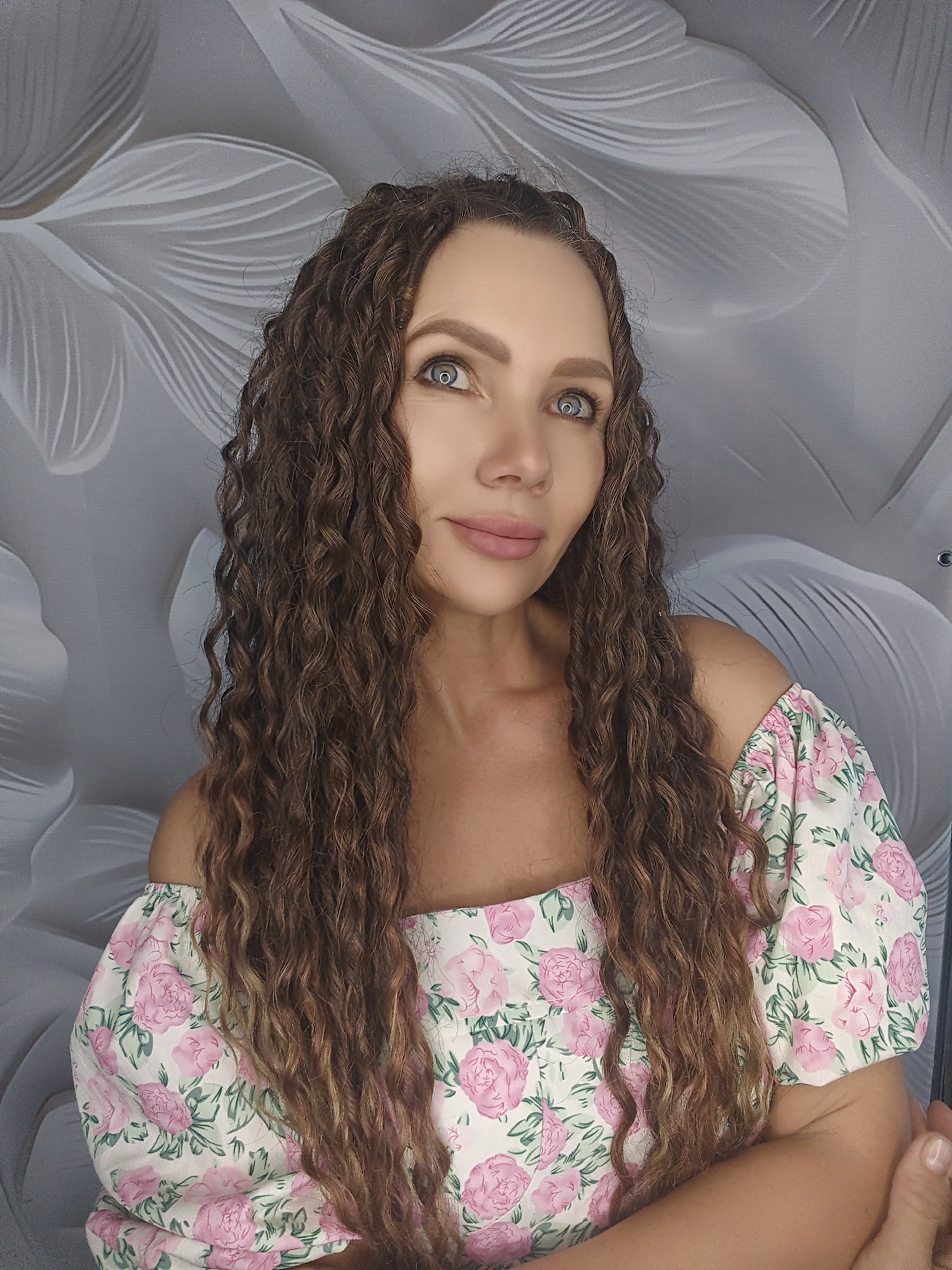 A portrait photograph shows a woman with long, curly brown hair and light blue eyes. She is wearing an off-the-shoulder dress with a white background and a pink floral pattern. Her head is tilted slightly to her left, and she is looking upwards and to her right. The background is a textured gray wall with a white, abstract floral design.