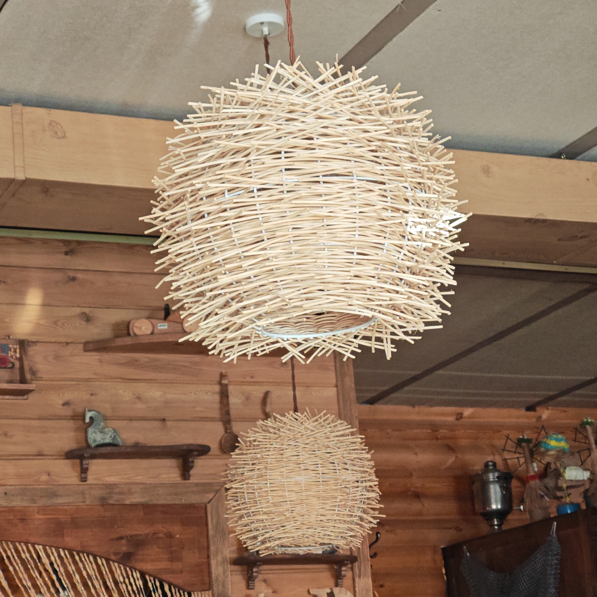 Two spherical lampshades made of woven light-colored wooden sticks hang from the ceiling of a room with wooden walls. The larger lampshade is suspended from a white ceiling mount with a brown cord. The smaller lampshade hangs below and slightly behind the larger one. The room has wooden walls and shelves with various decorative objects. Novo-Nikol'skoe, Russia, is not evident in the image.