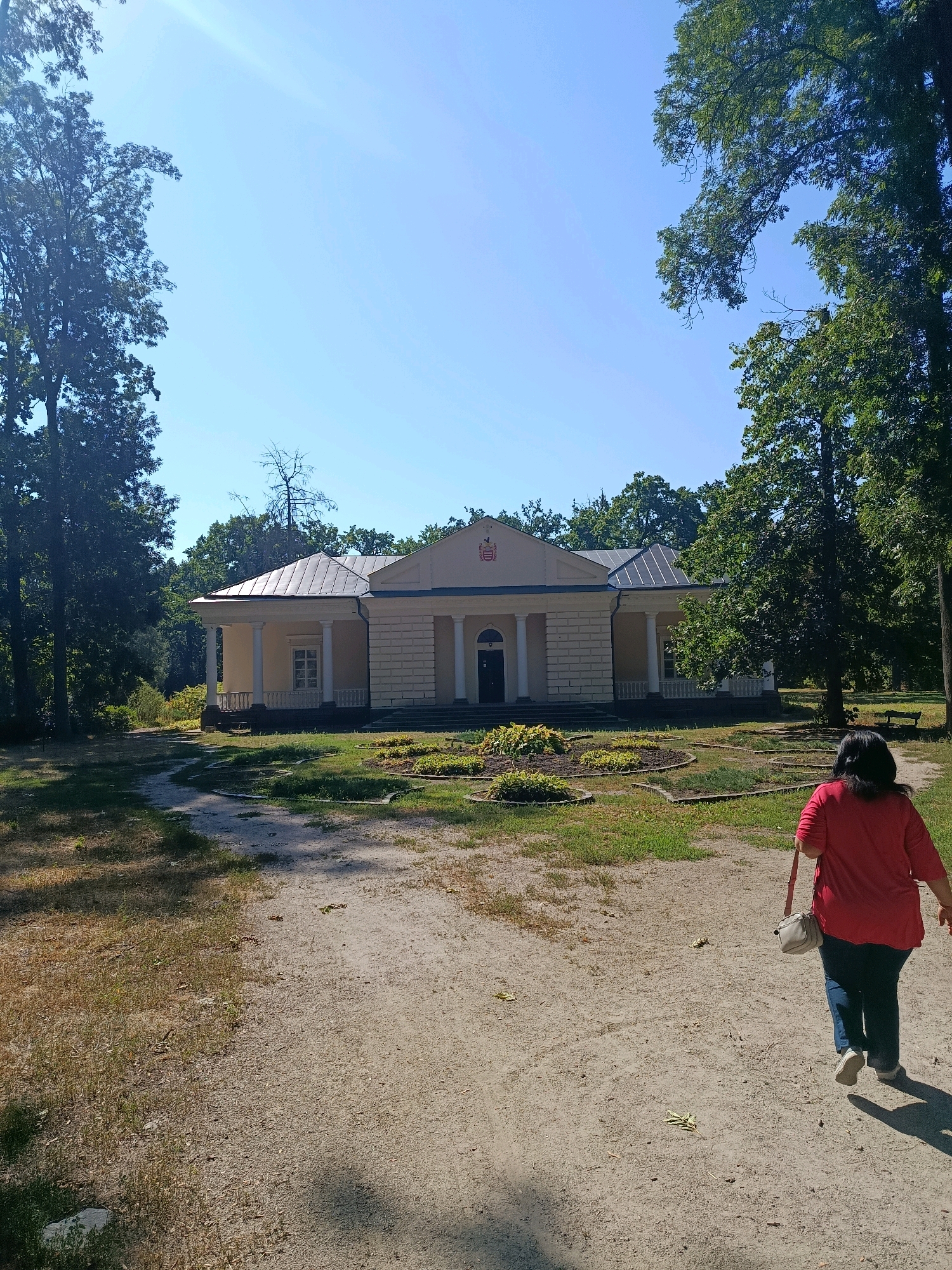 The image depicts the park zone of the Olexander's Garden in Bila Tserkva, Ukraine. In the foreground, a woman wearing a red shirt and blue jeans walks away from the camera on a dirt path. She carries a light-colored crossbody bag. Behind her, a light-colored, single-story building with a gray roof and columns is visible. The building is surrounded by green trees and a landscaped garden area. The sky is clear and blue, with a slight lens flare in the upper left corner. The overall scene is that of a sunny day in a park.